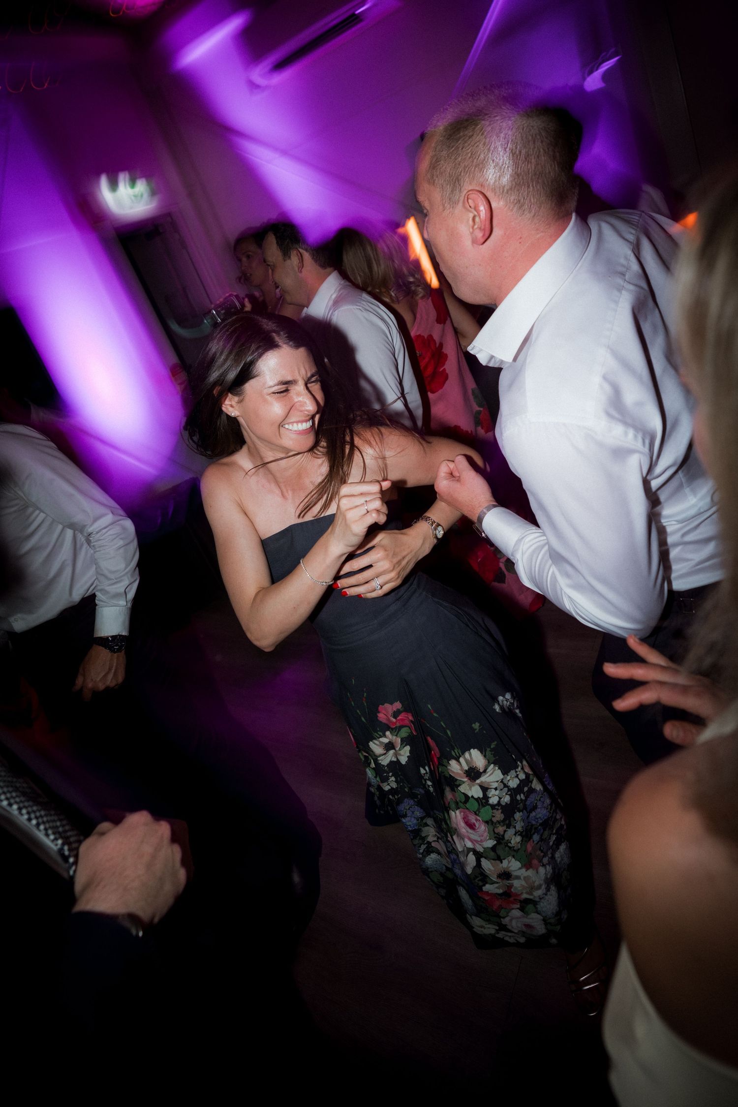 People dancing and celebrating on a dance floor illuminated by purple lighting at a nighttime event.