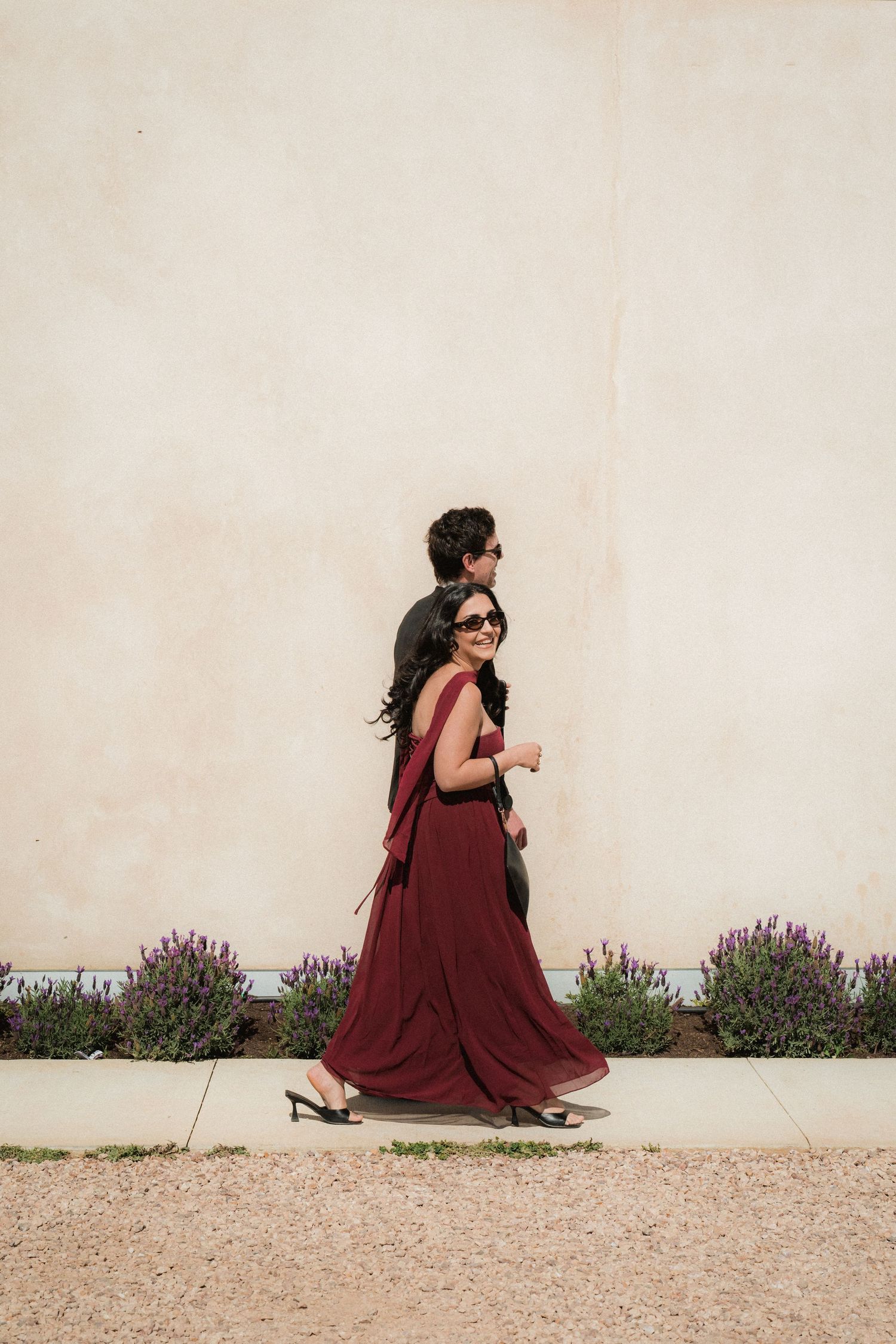 A smiling woman in a flowing burgundy dress and sunglasses walks along a sunlit wall with a man behind her and lavender plants.