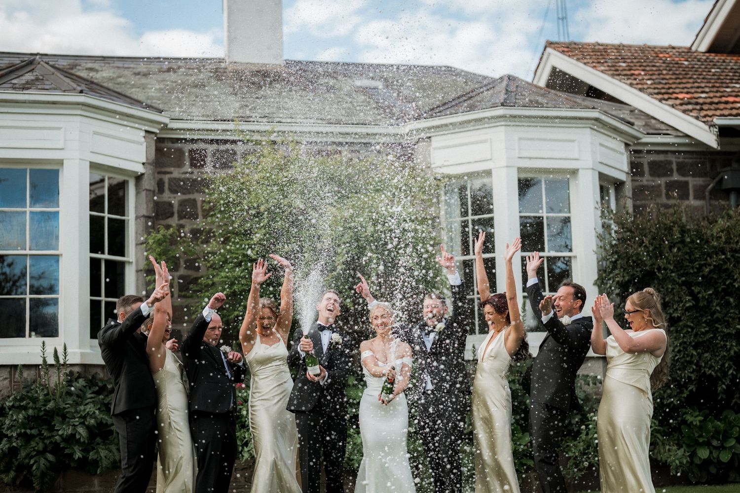 Wedding party celebrating with champagne spray outside elegant stone building.
