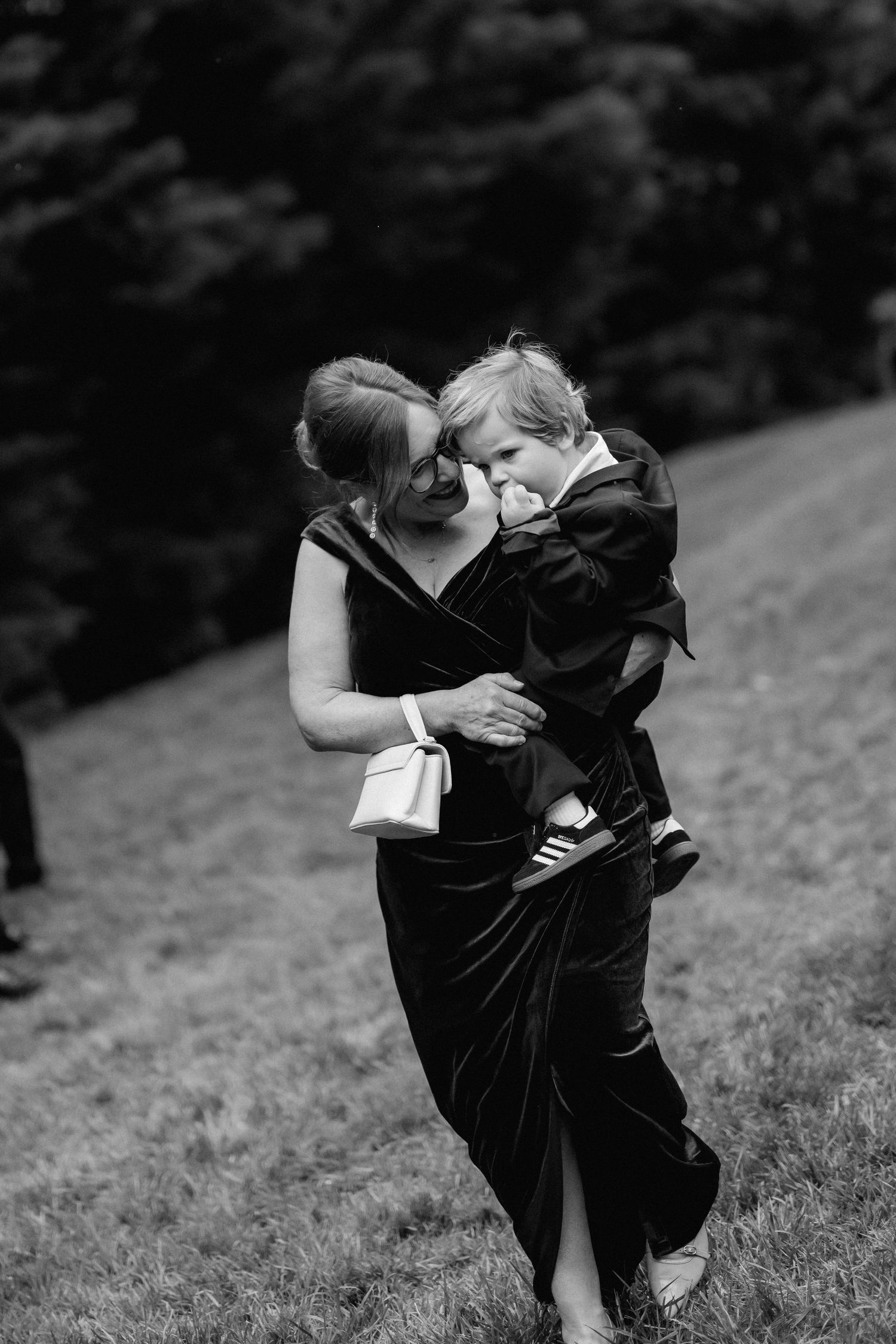 A tender moment captured in black and white as a mother in a velvet dress embraces a small child on a grassy slope.