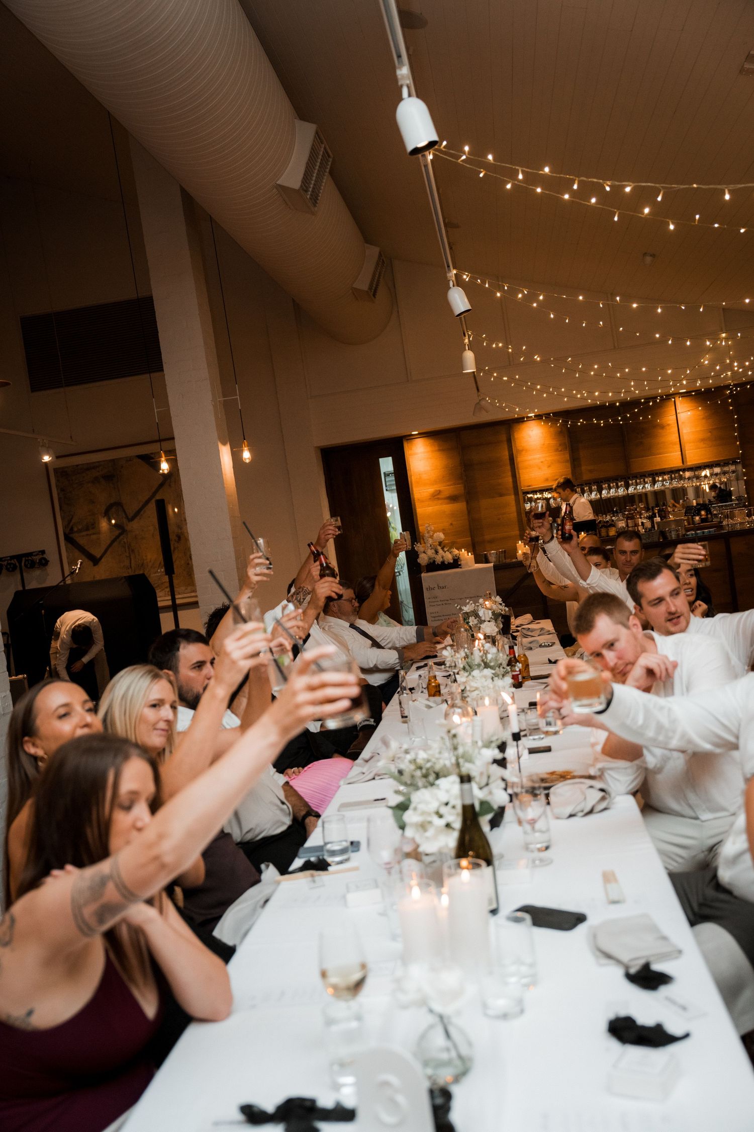 Wedding guests raise their glasses in celebration at a long reception table decorated with string lights overhead.