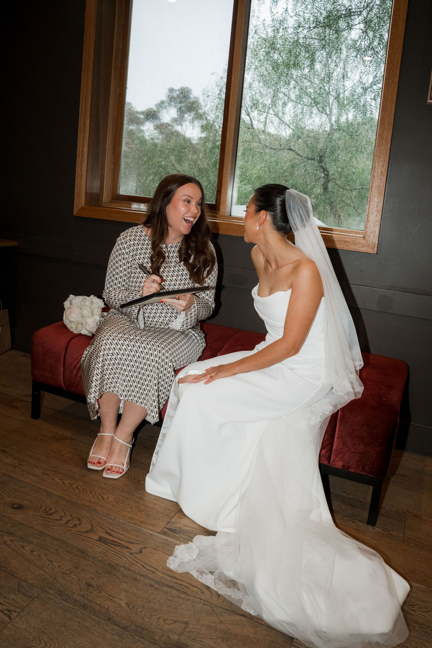 A bride in a flowing white gown sits on a wooden floor during wedding preparations.