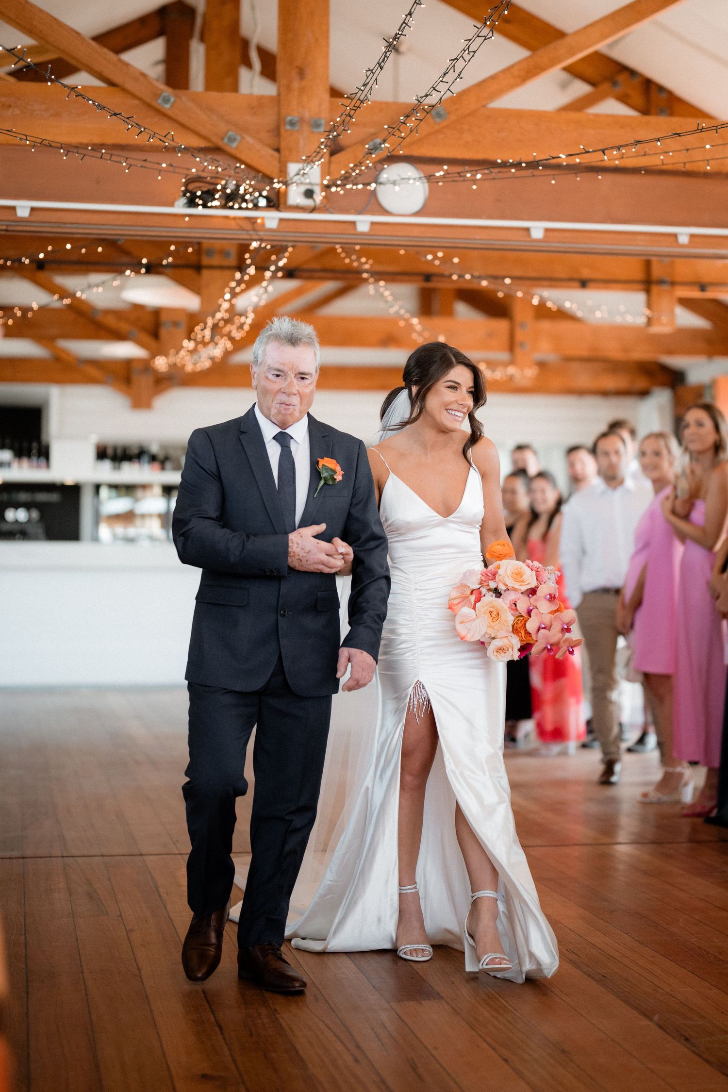 Wedding processional in rustic venue with wooden beams and string lights overhead.