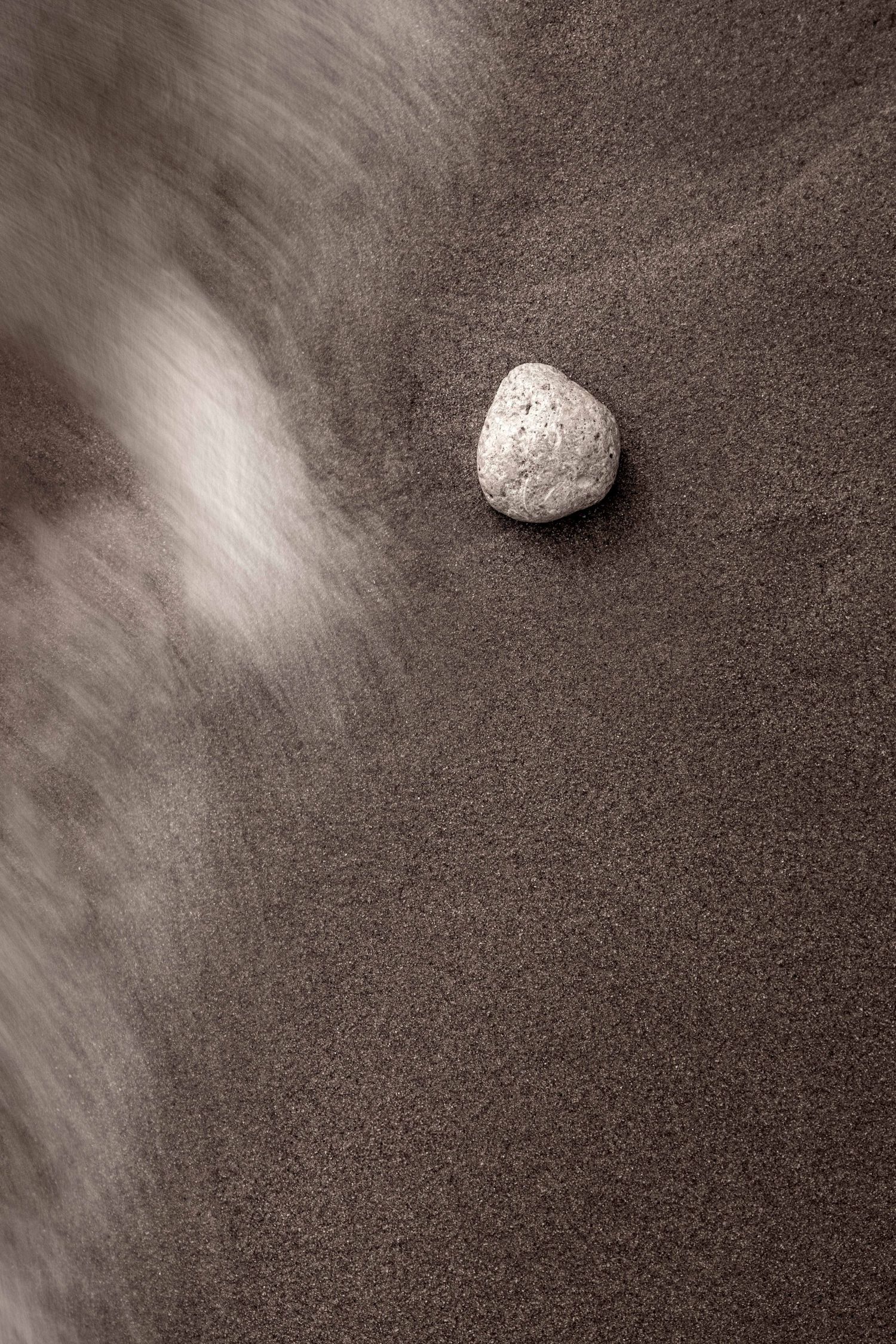 Abstract fine art photograph of a single pebble on dark sand with a wave washing past, Algarve beach photography
