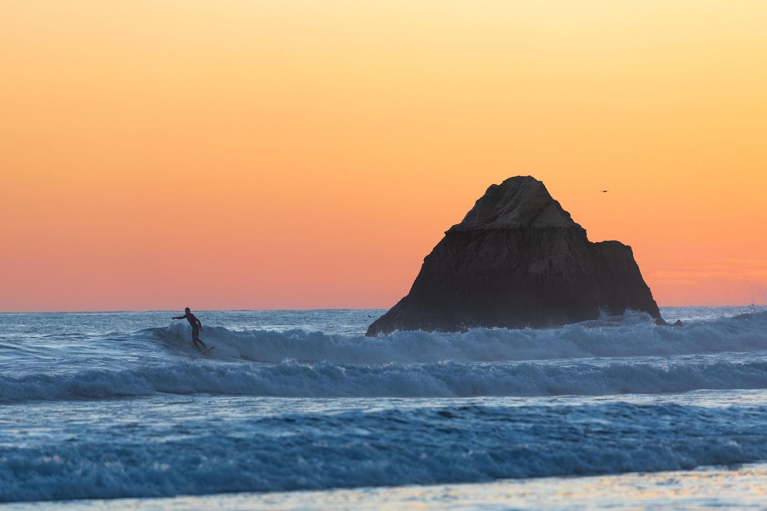 Surfer in the waves at sunset beside a dramatic sea stack at Portimão beach, Algarve, Portugal