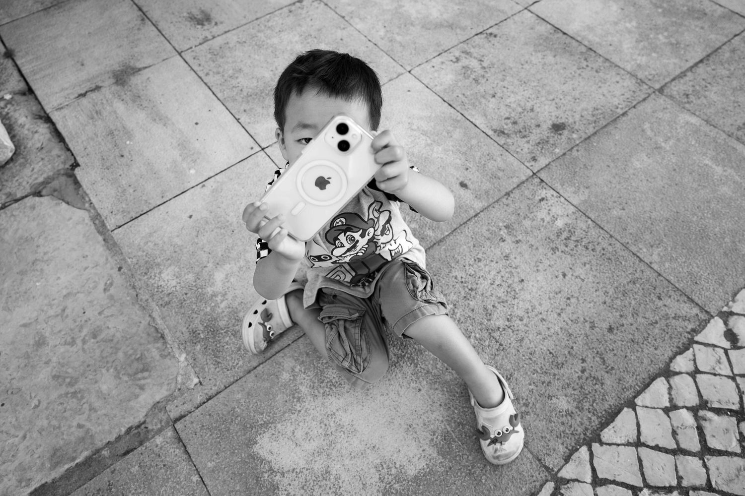 Black and white candid street photograph of a young boy holding a phone camera, documentary photography by Anthony West
