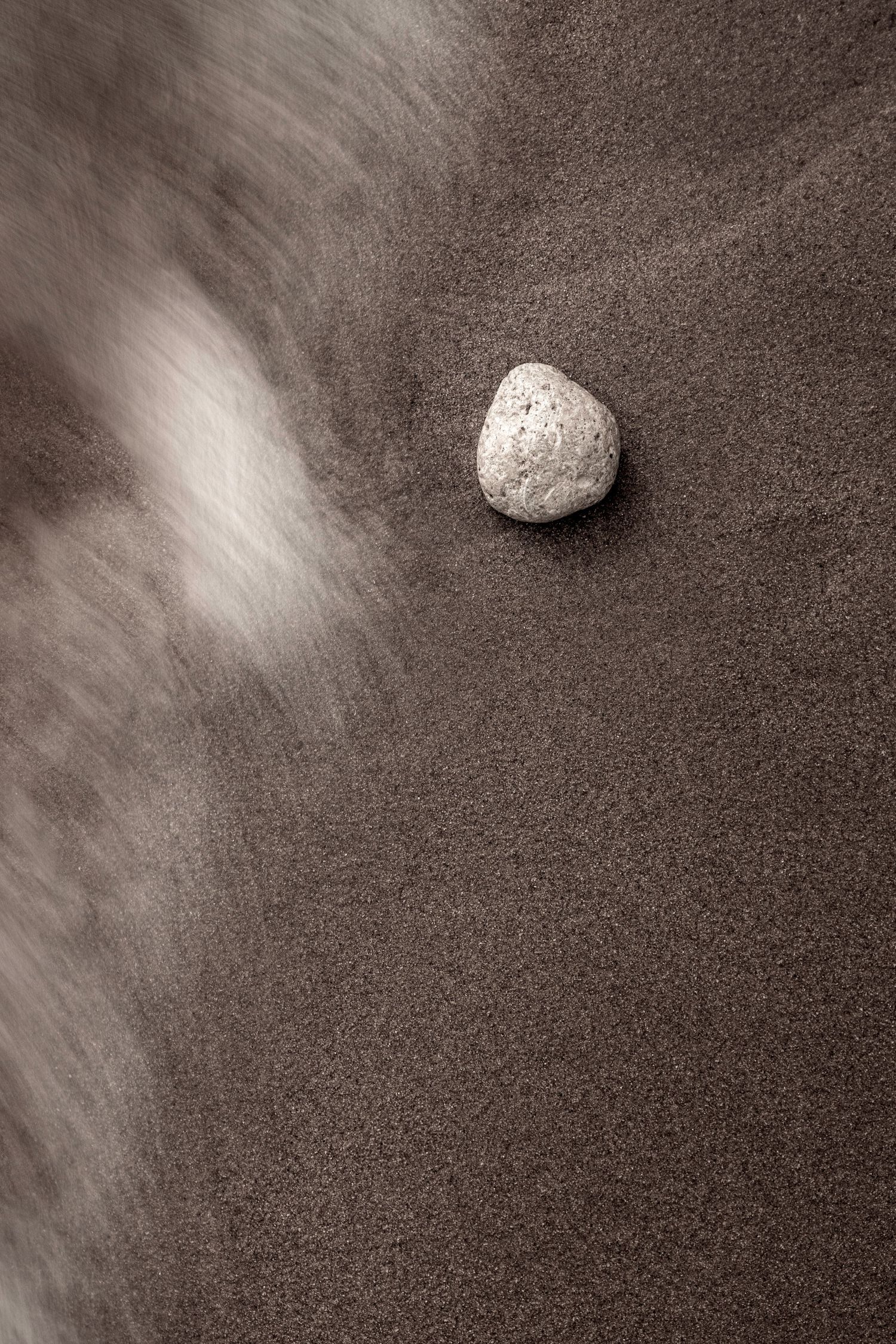 Abstract fine art photograph of a single pebble on dark sand with a wave washing past, Algarve beach photography