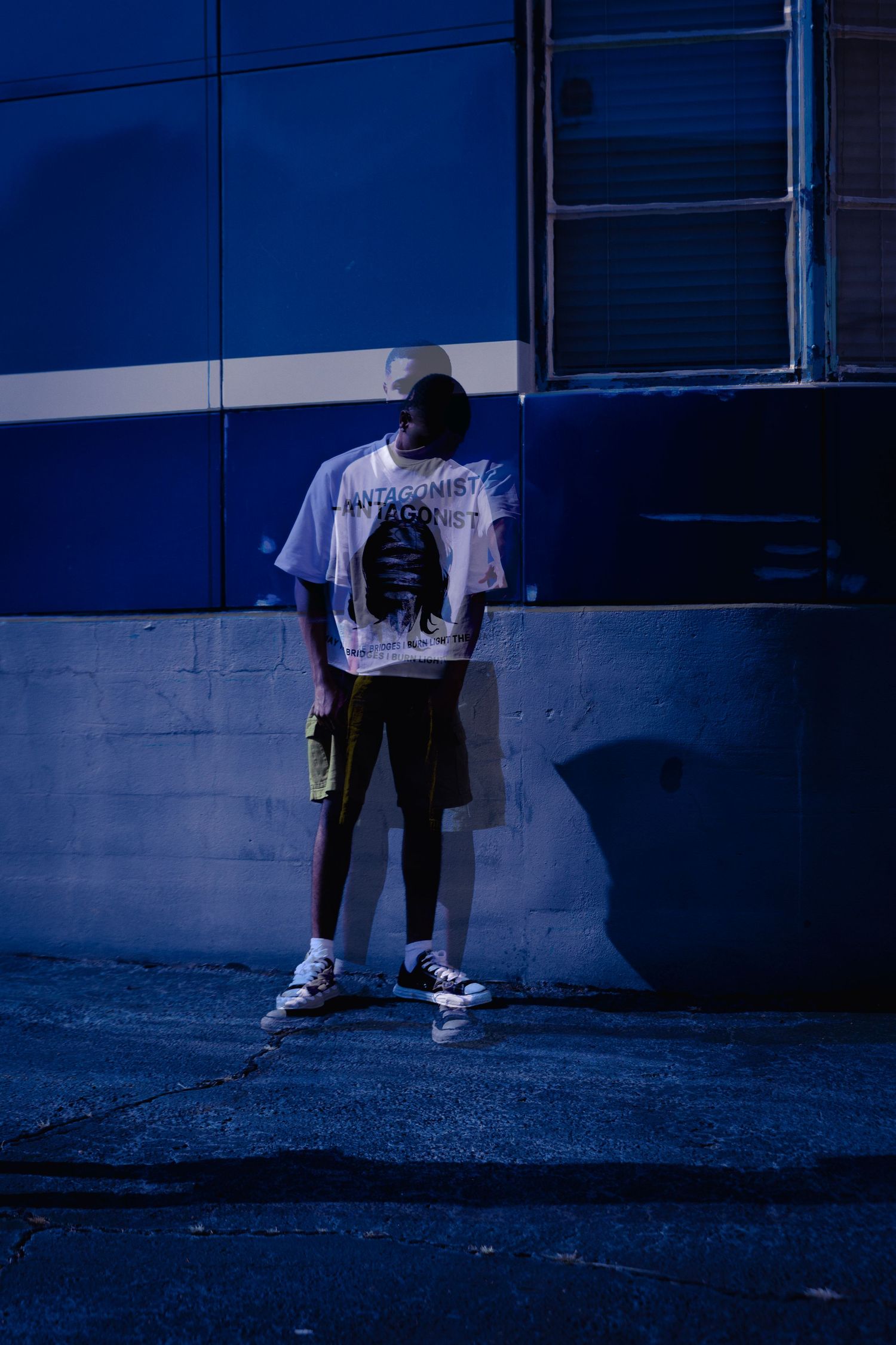 Person in white graphic tee and shorts leaning against blue building wall at night.