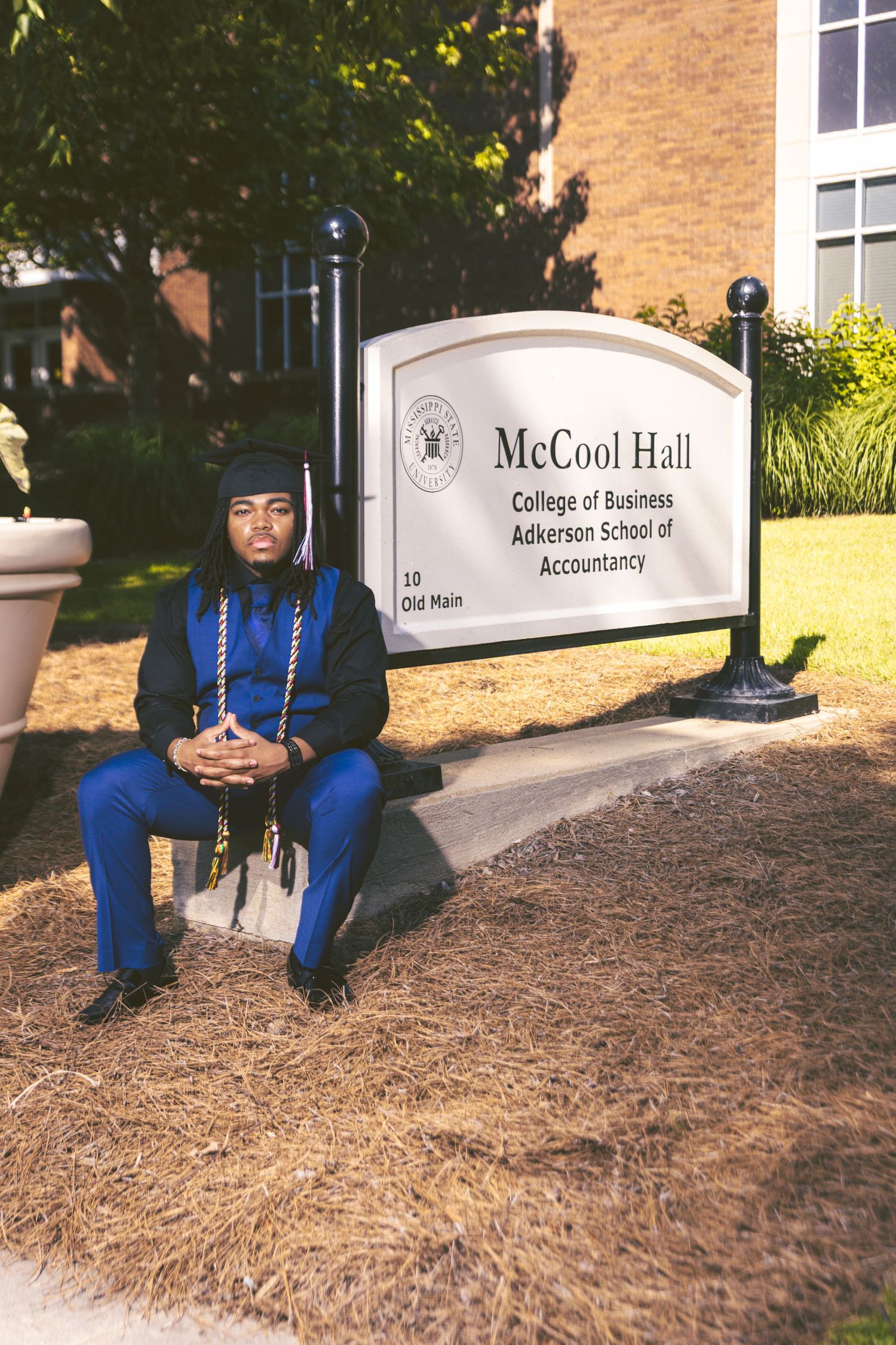 Black male graduate in blue regalia sits beside McCool Hall College of Business sign.