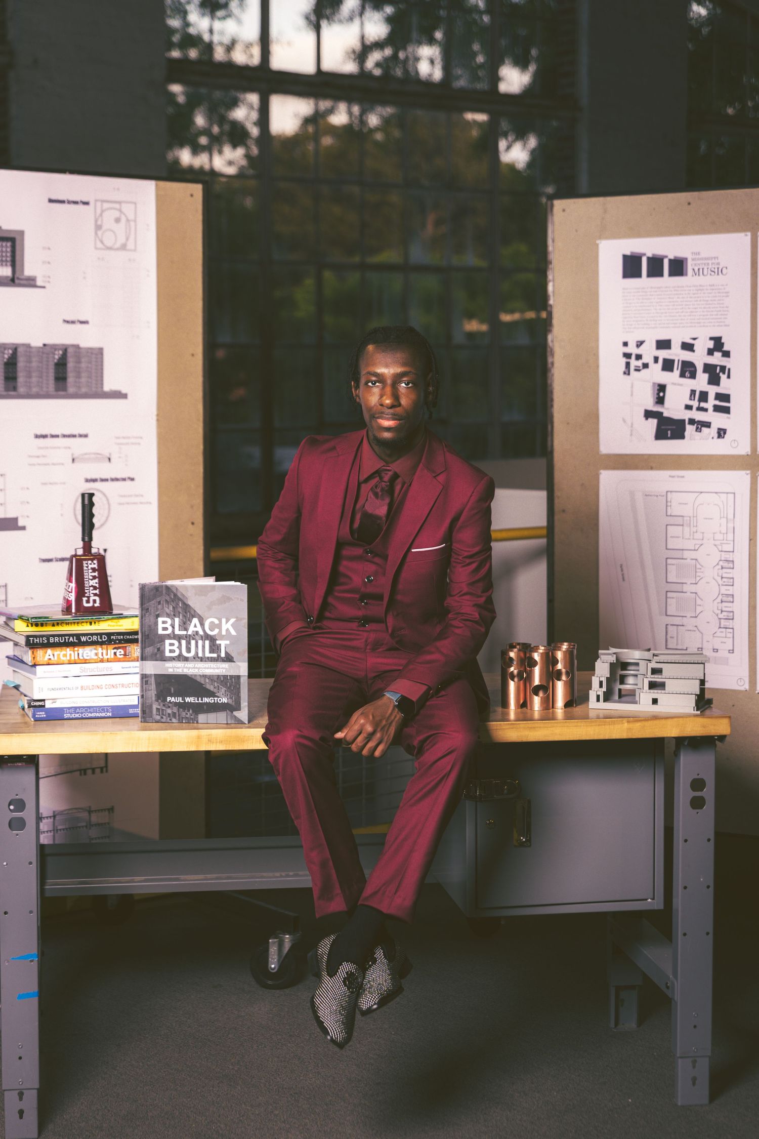 Black male graduate in maroon suit sits at desk surrounded by architecture books and models.