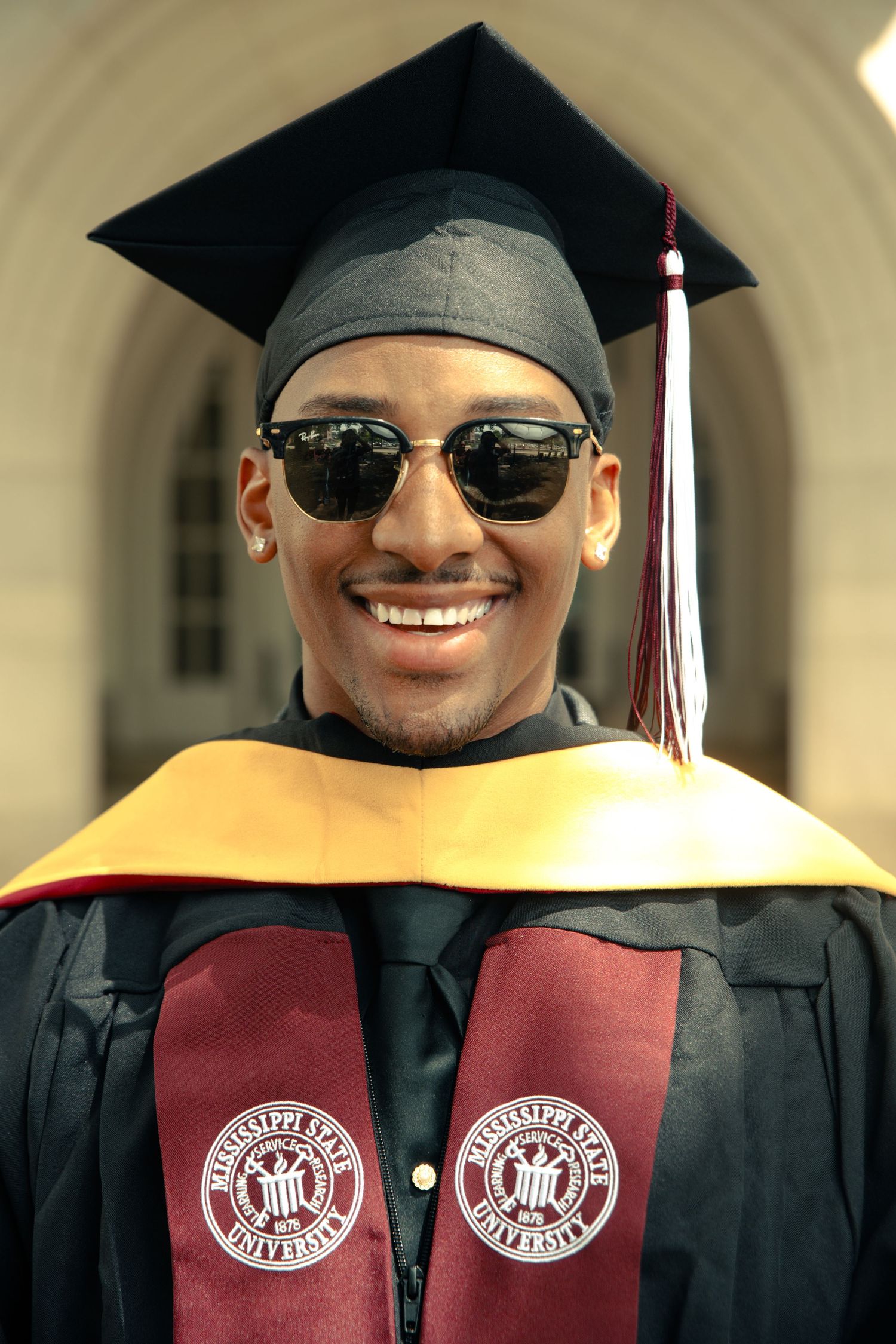 Smiling Black male graduate in Mississippi State cap and gown wearing sunglasses outdoors.