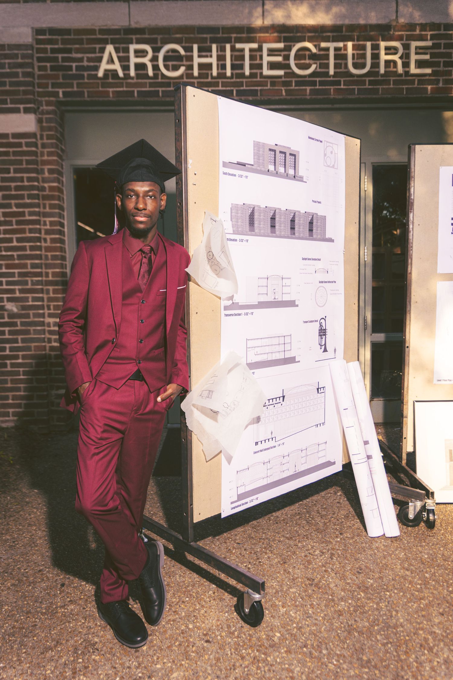 Black male architecture graduate in maroon suit stands beside architectural drawings display.