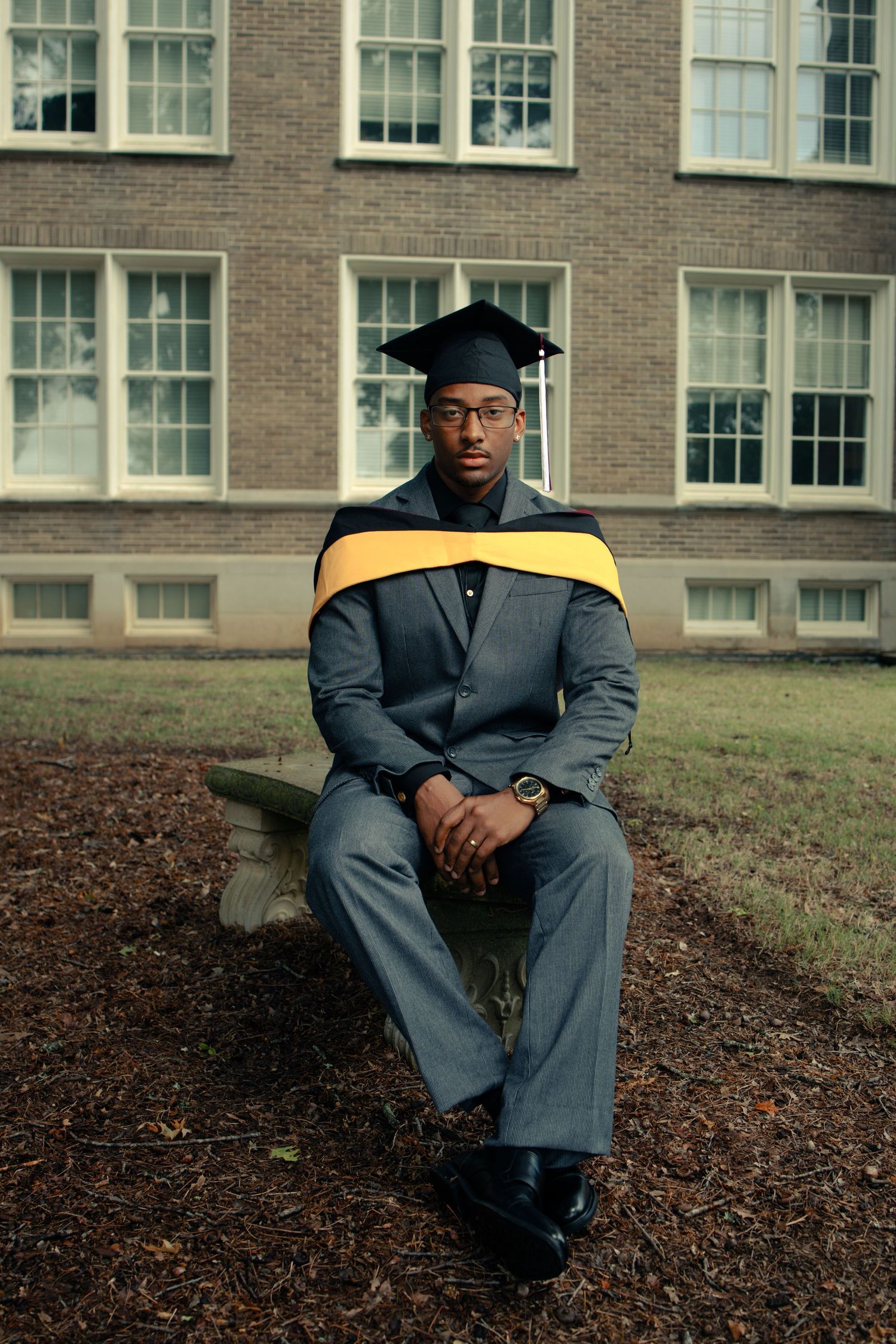 Black male graduate in gray suit with gold hood sits on bench outside brick building.