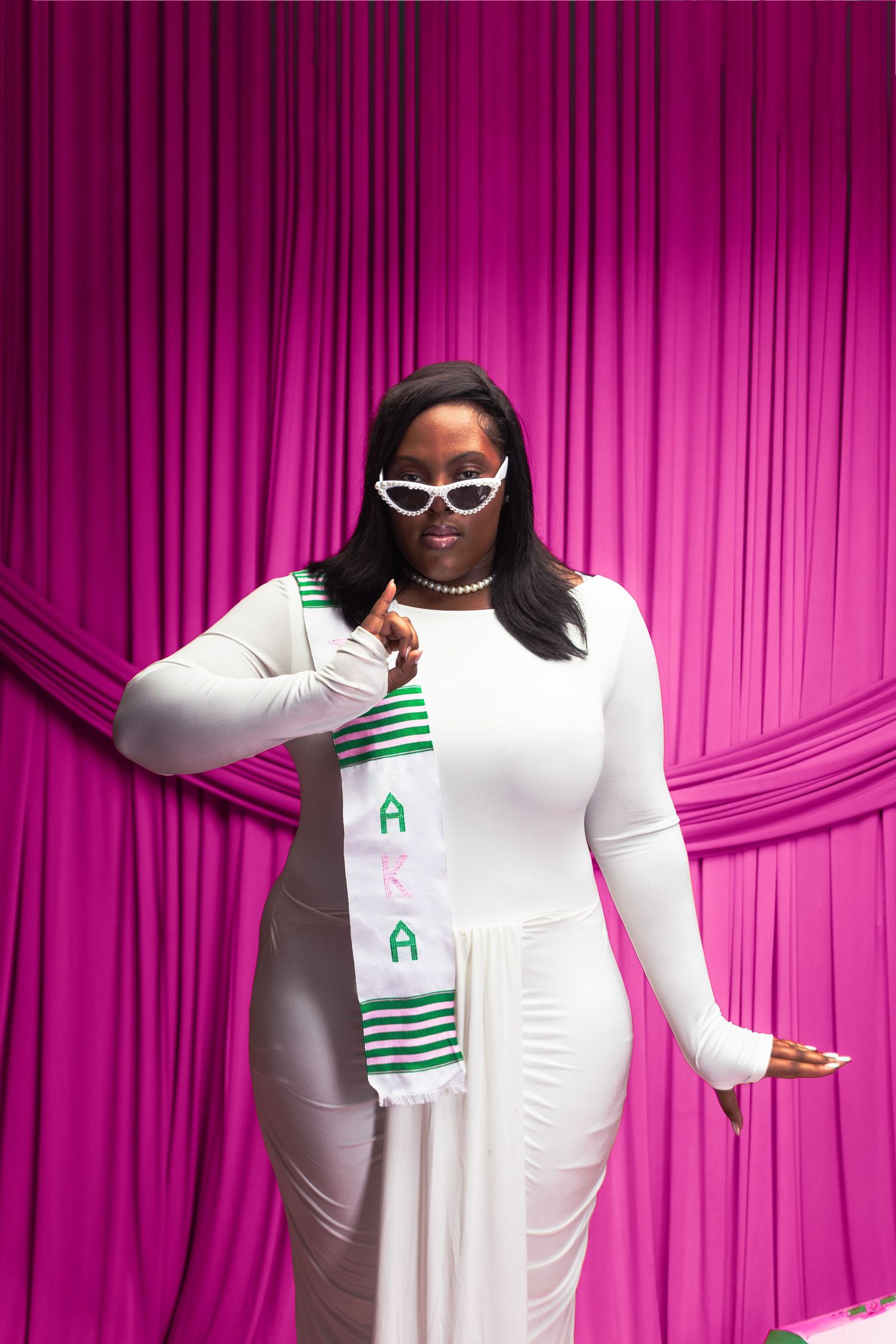 Black woman in white dress with AKA sash poses confidently before pink curtain backdrop.
