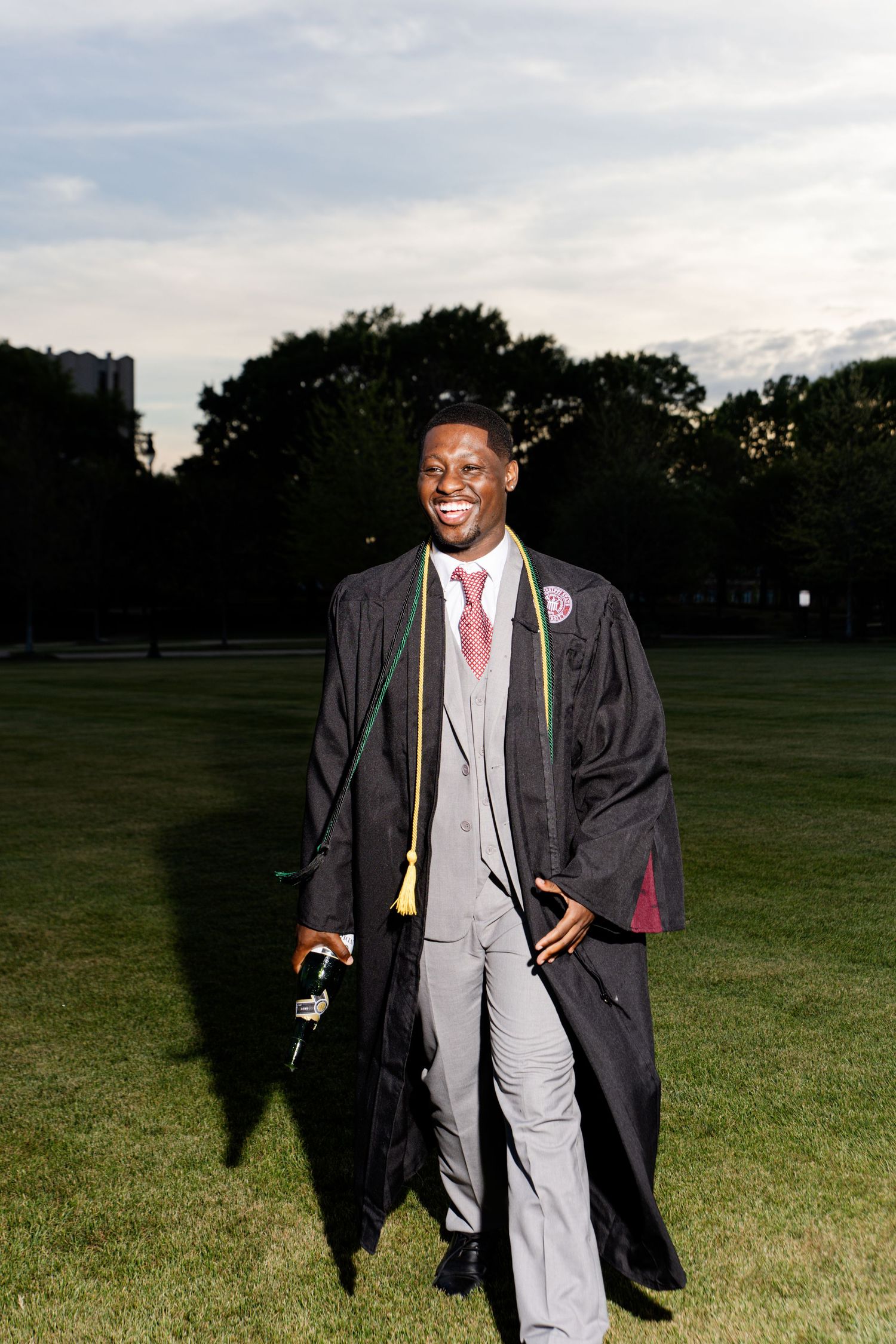 Smiling graduate in cap and gown with colorful cords walks across green lawn.
