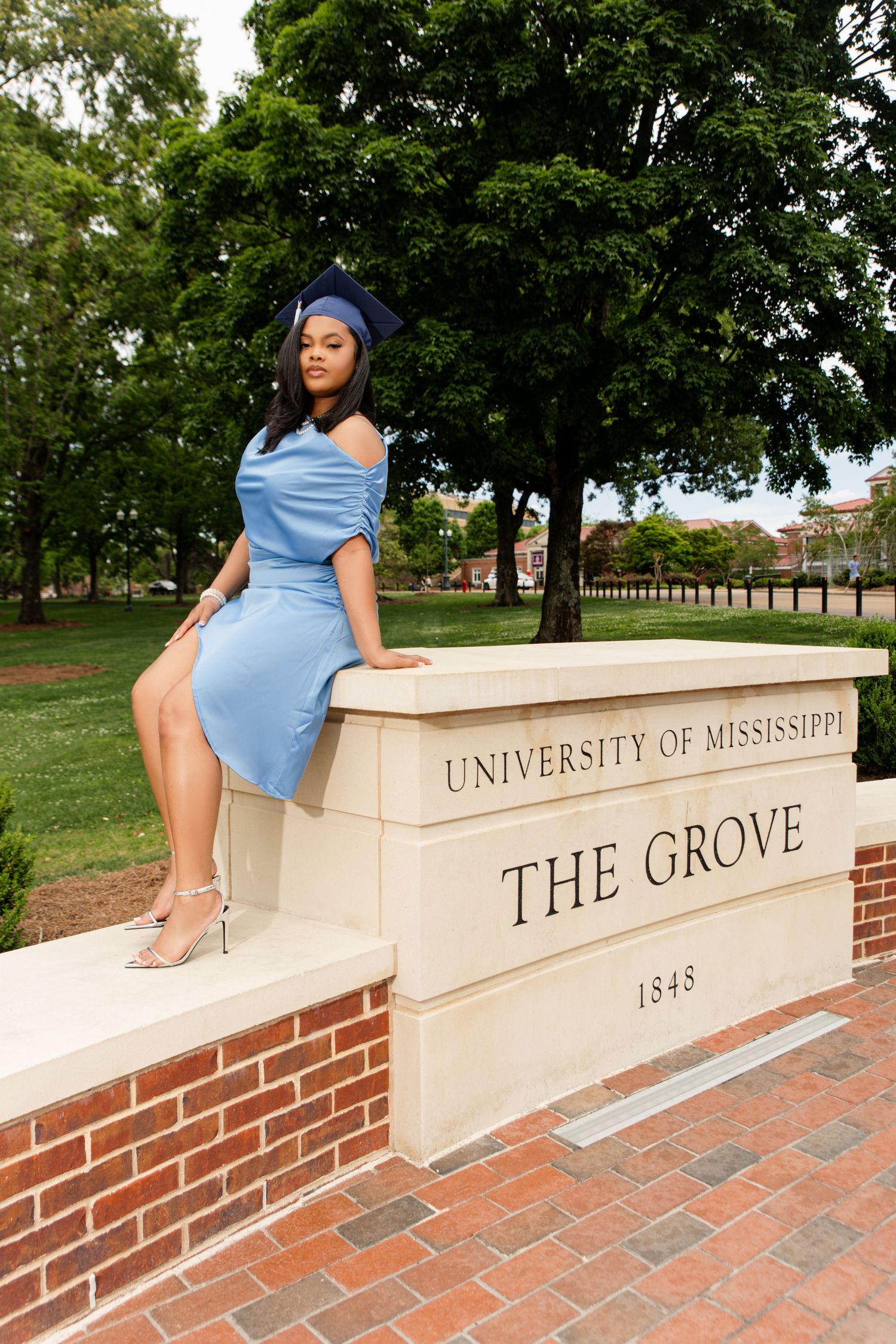 Graduate in blue dress and cap sits on University of Mississippi Grove sign.