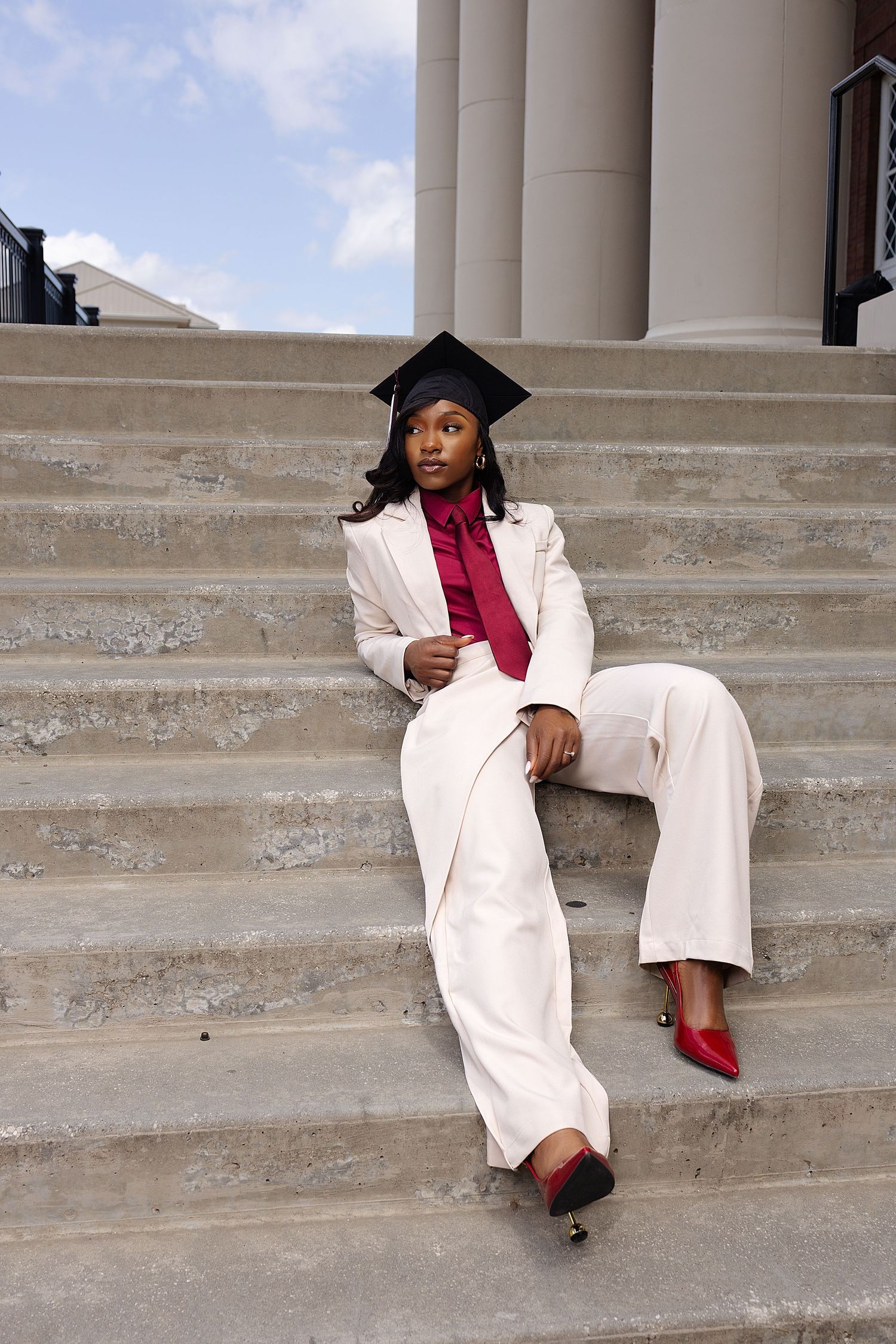 Black woman in white suit and graduation cap poses on university steps in red heels.