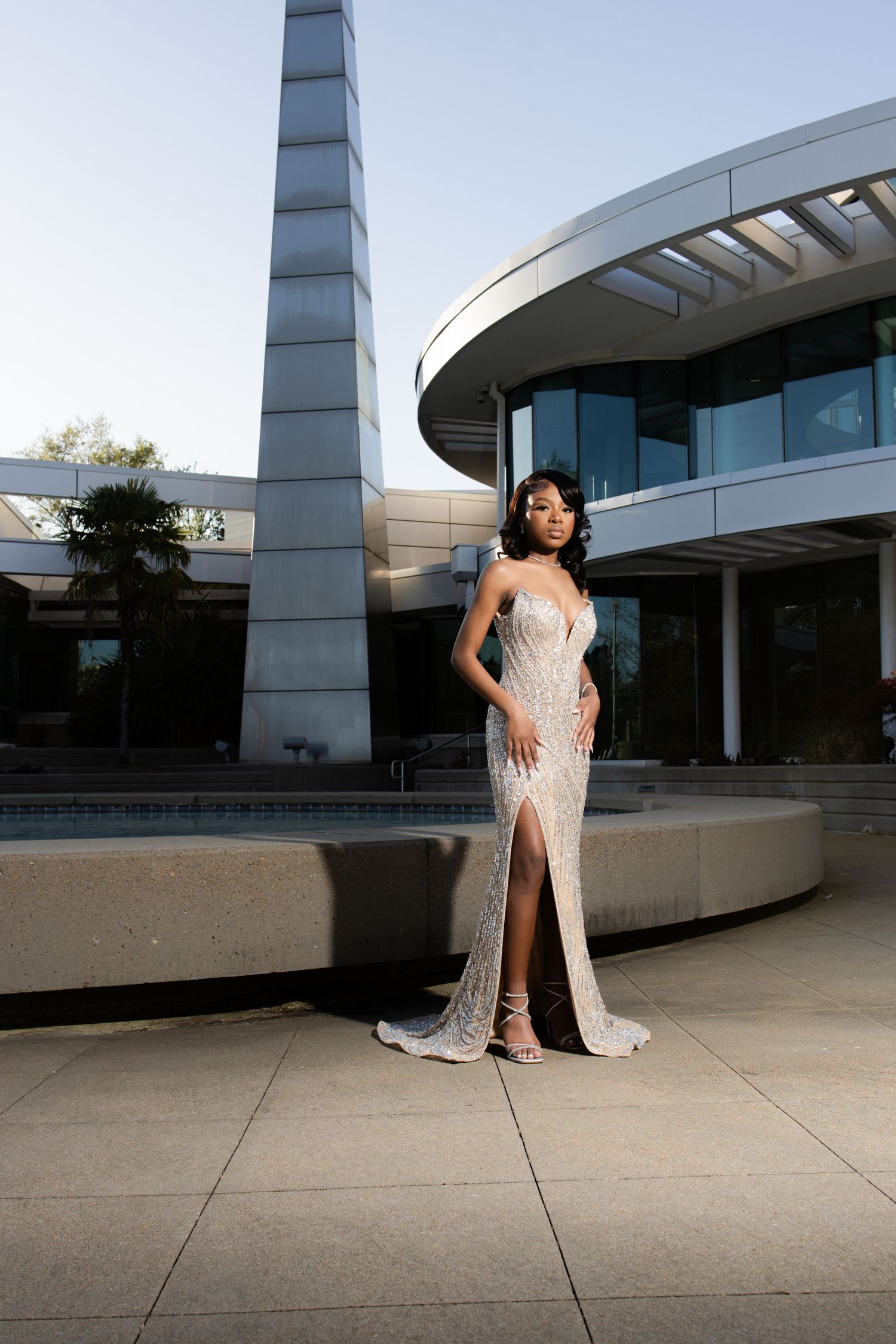 Woman in elegant beaded gown poses outside modern architectural building at dusk.