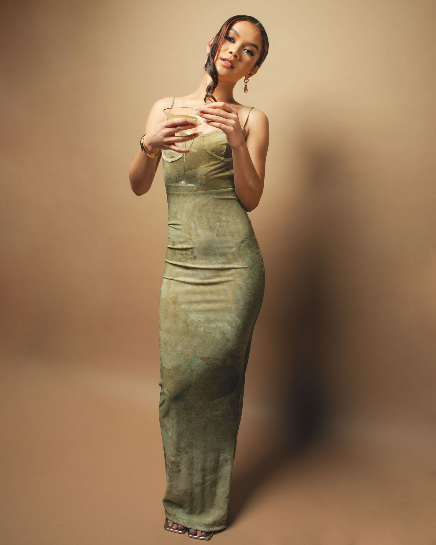 Woman posing in elegant sage green spaghetti strap maxi dress against a warm beige background.