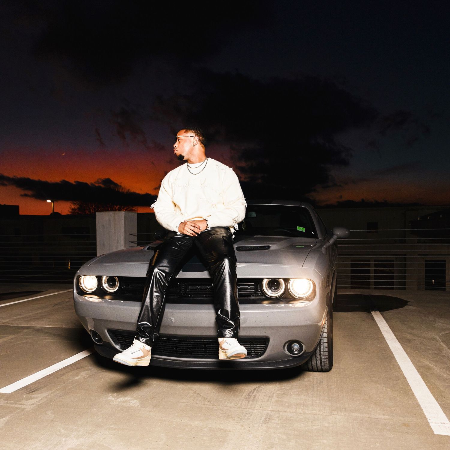 Man in white hoodie sits on gray Dodge Challenger during colorful sunset.