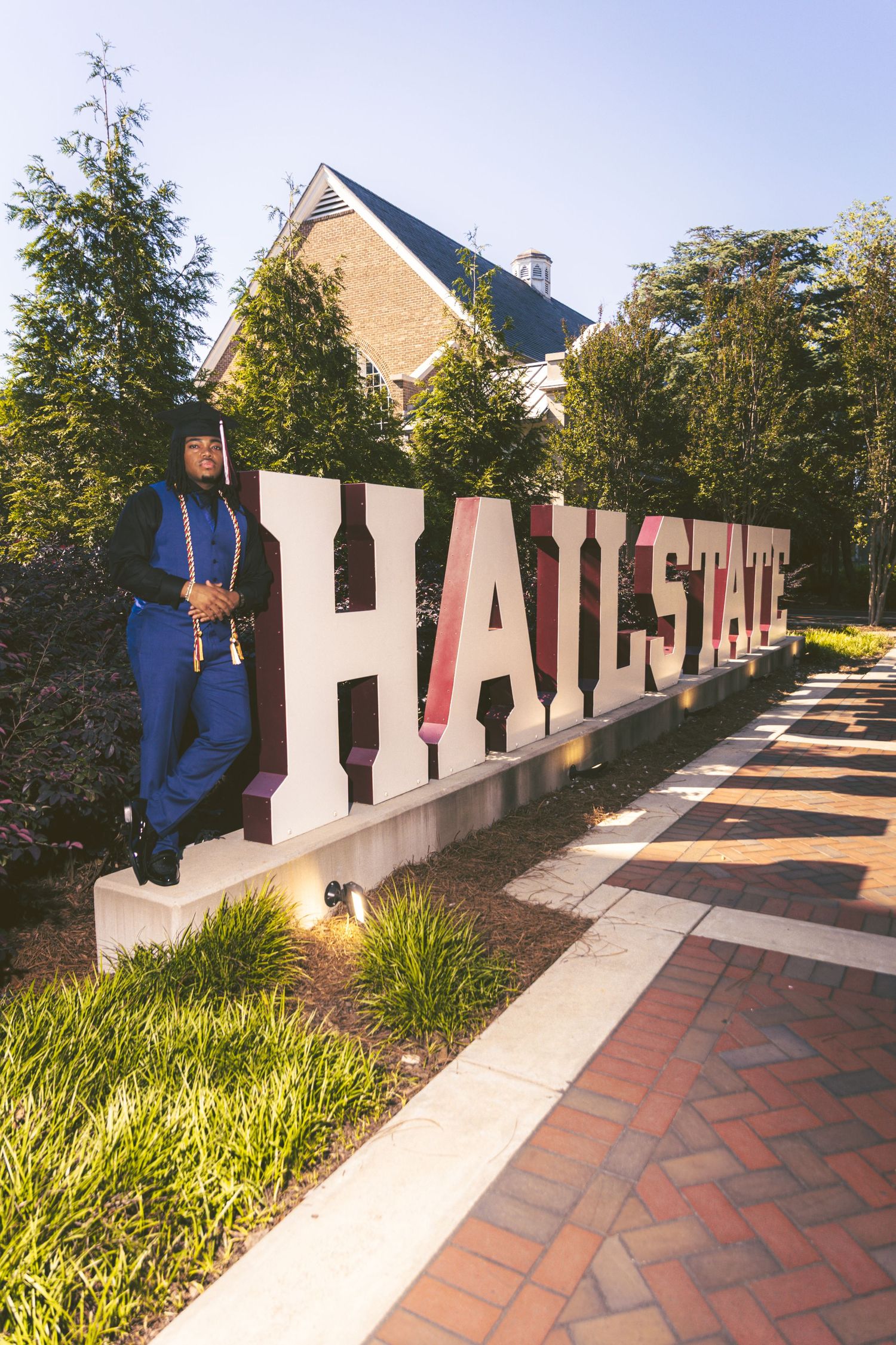 Black female graduate in blue cap and gown leans against Hail State campus sign.
