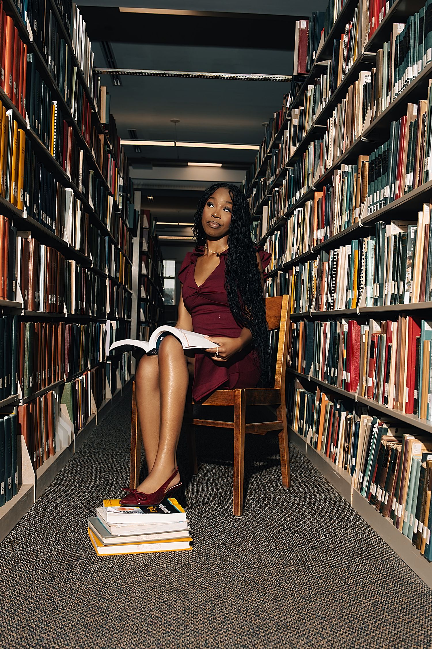 Woman in burgundy dress reads book while seated in university library aisle.