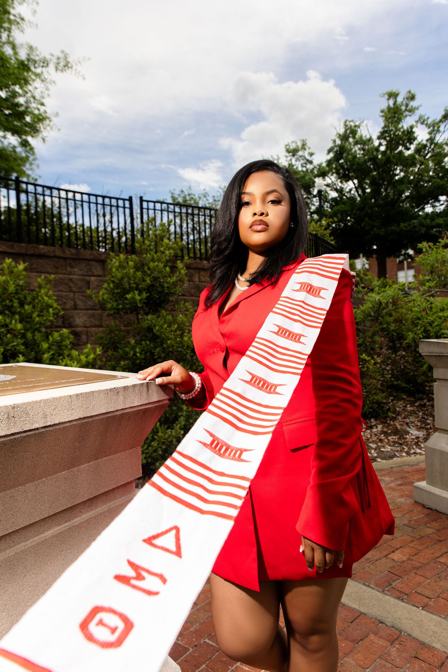 Woman in red blazer dress wearing Theta Delta Chi graduation sash outdoors.