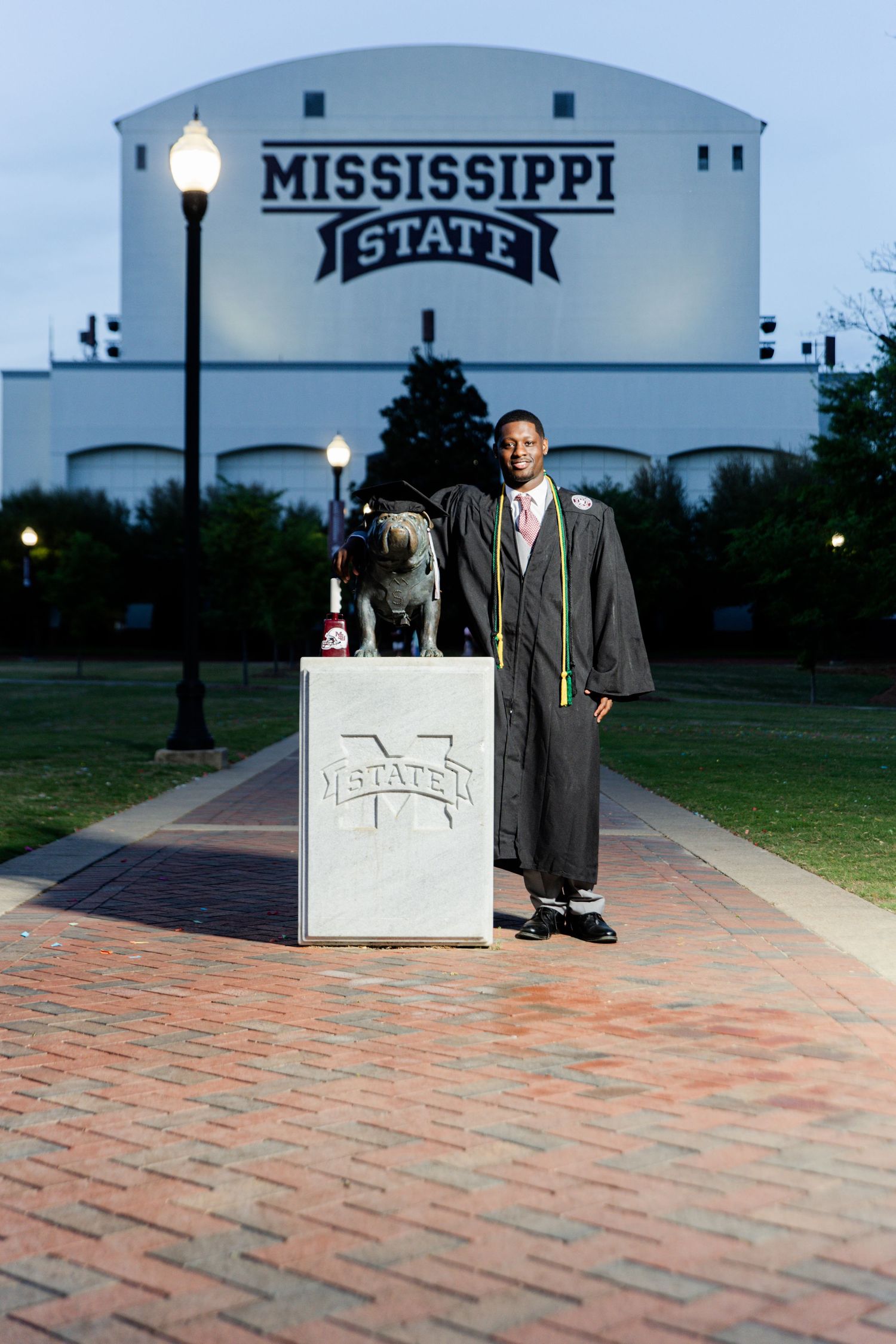 Graduate in cap and gown poses by Mississippi State Bulldog mascot statue.