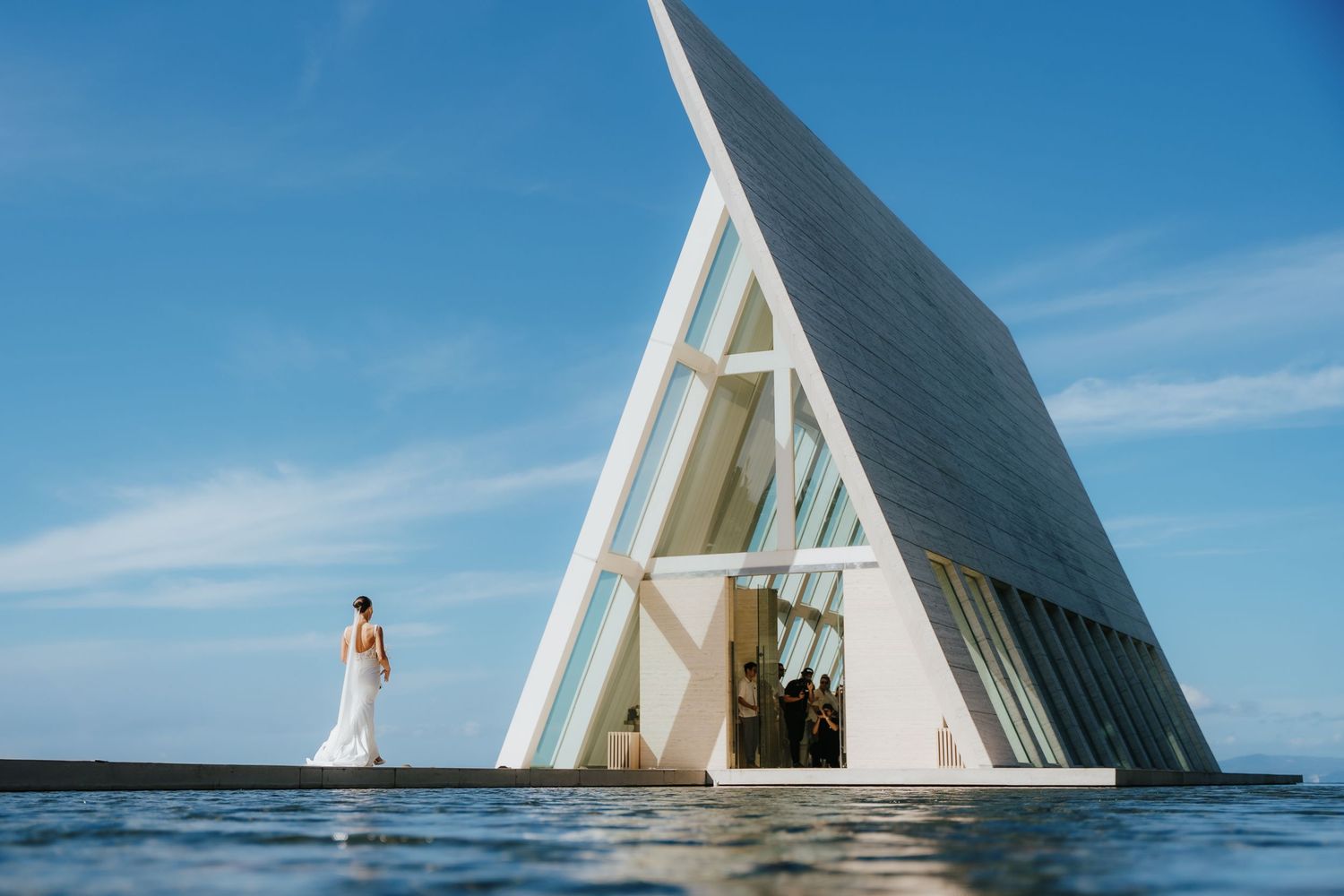 A modern triangular white chapel at Conrad Bali with a dramatic slanted roof stands in shallow water against a bright blue sky.