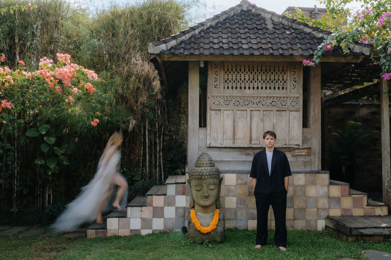 Couple poses near ornate Balinese wooden shrine with Buddha statue and marigold garland in lush tropical garden.