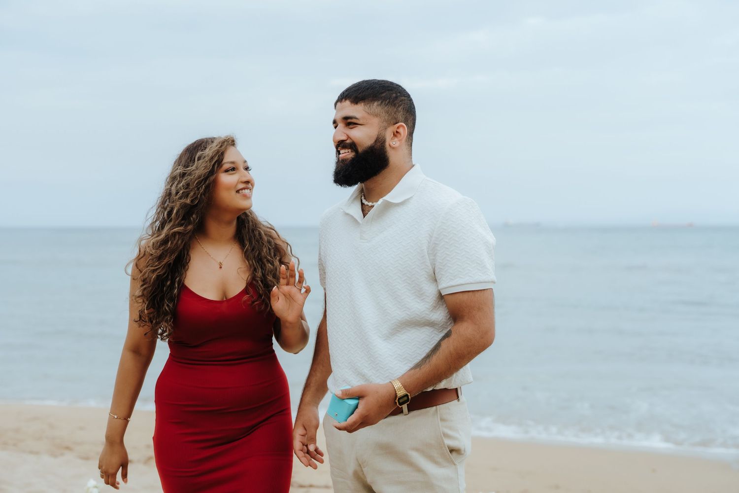 A couple in red and white outfits poses together during a romantic beach photoshoot at sunset.
