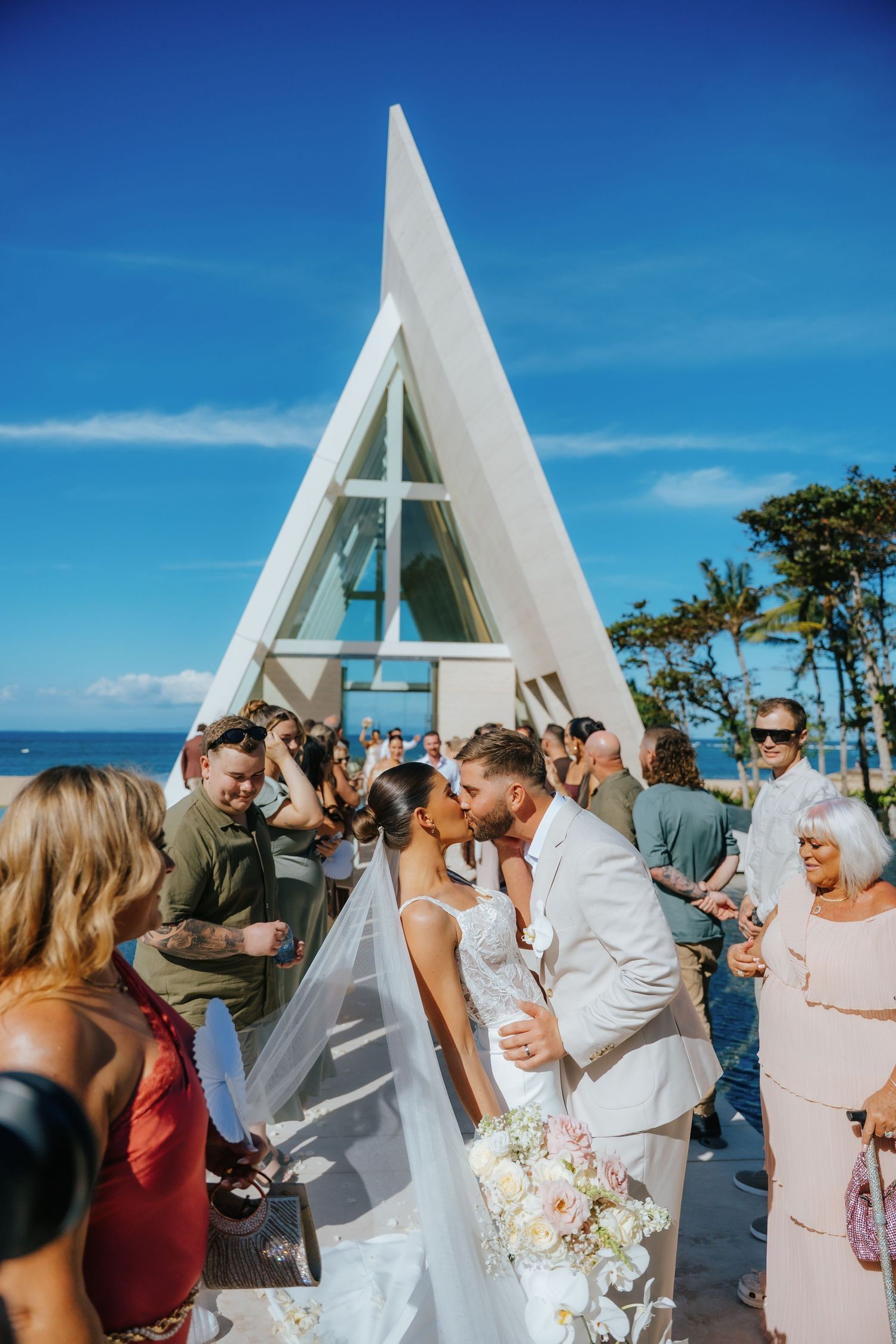 Bride and groom share a kiss at outdoor A-frame chapel wedding ceremony with ocean backdrop and guests celebrating.