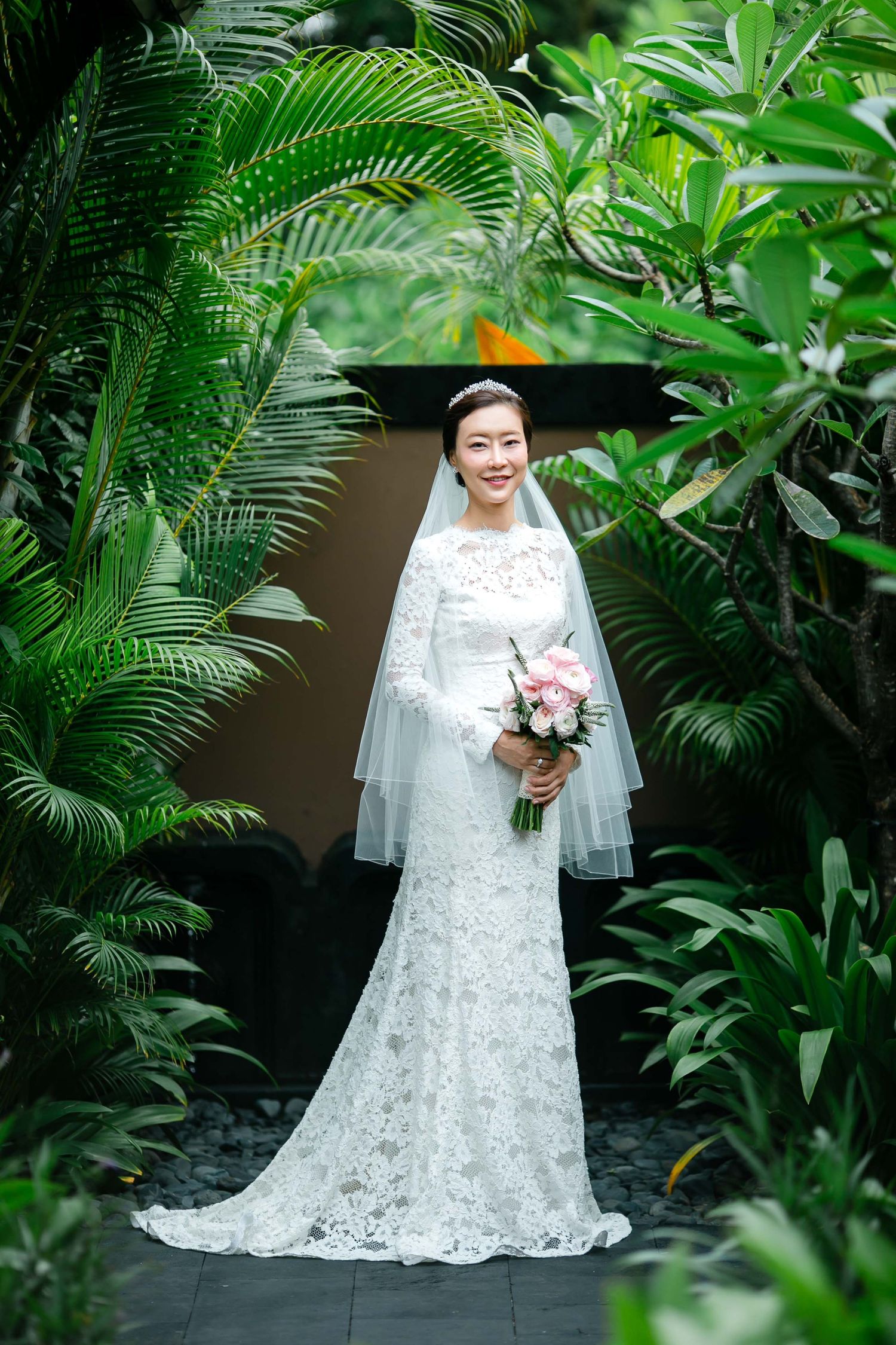 Smiling Asian bride in lace wedding gown and veil holds pink bouquet surrounded by lush tropical greenery.