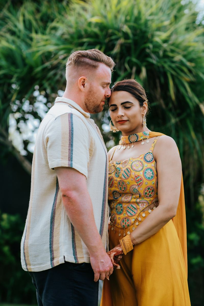 A couple in a romantic outdoor photoshoot wearing a yellow dress and white shirt against a tropical palm background.