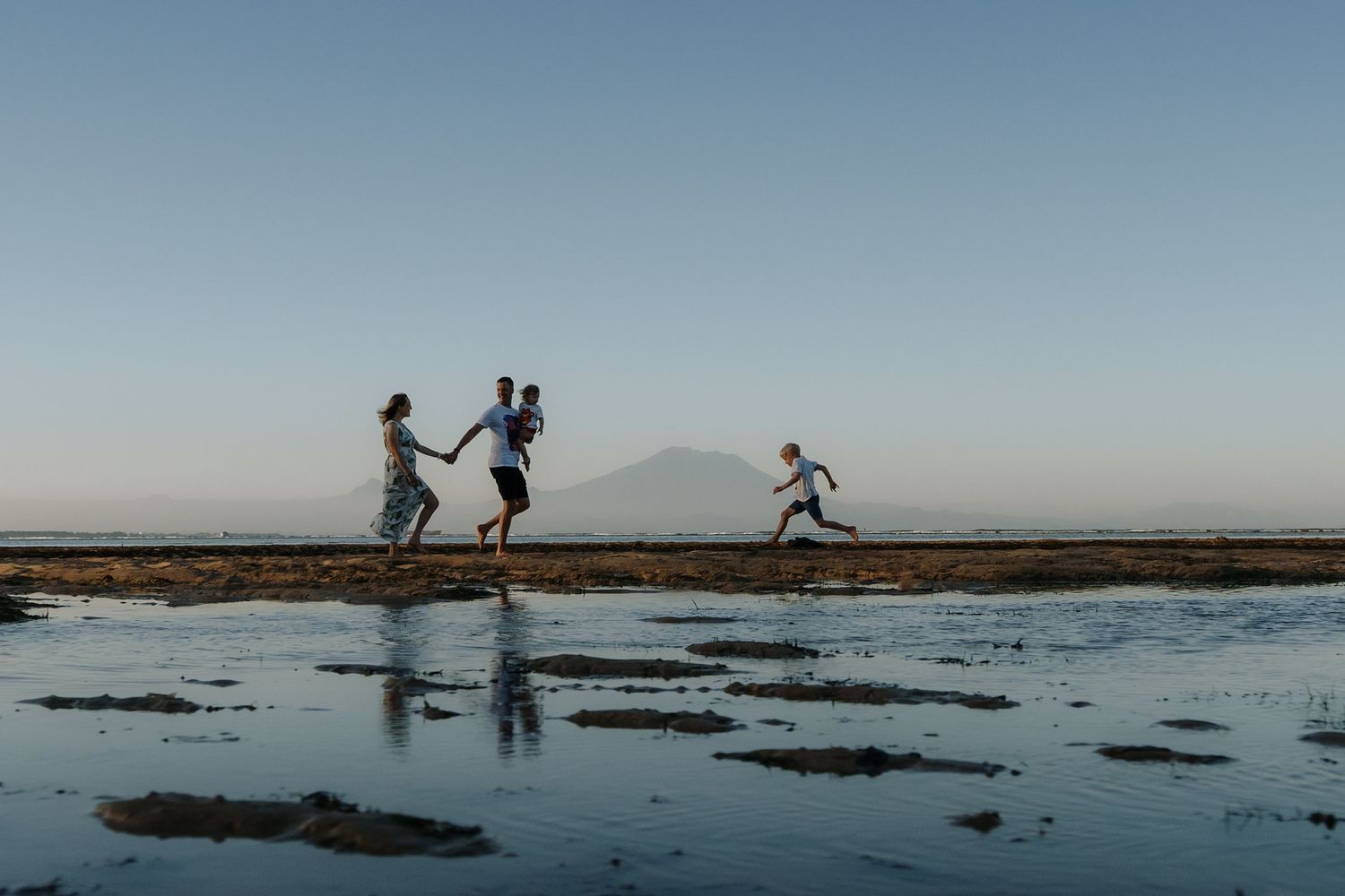 Family running and playing on a reflective beach at sunset with a mountain silhouette in the background.