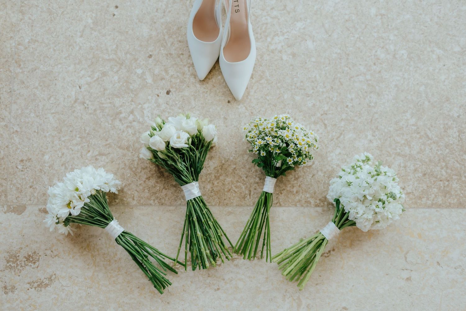 Four white wedding bouquets arranged on stone floor with bridal heels in background.