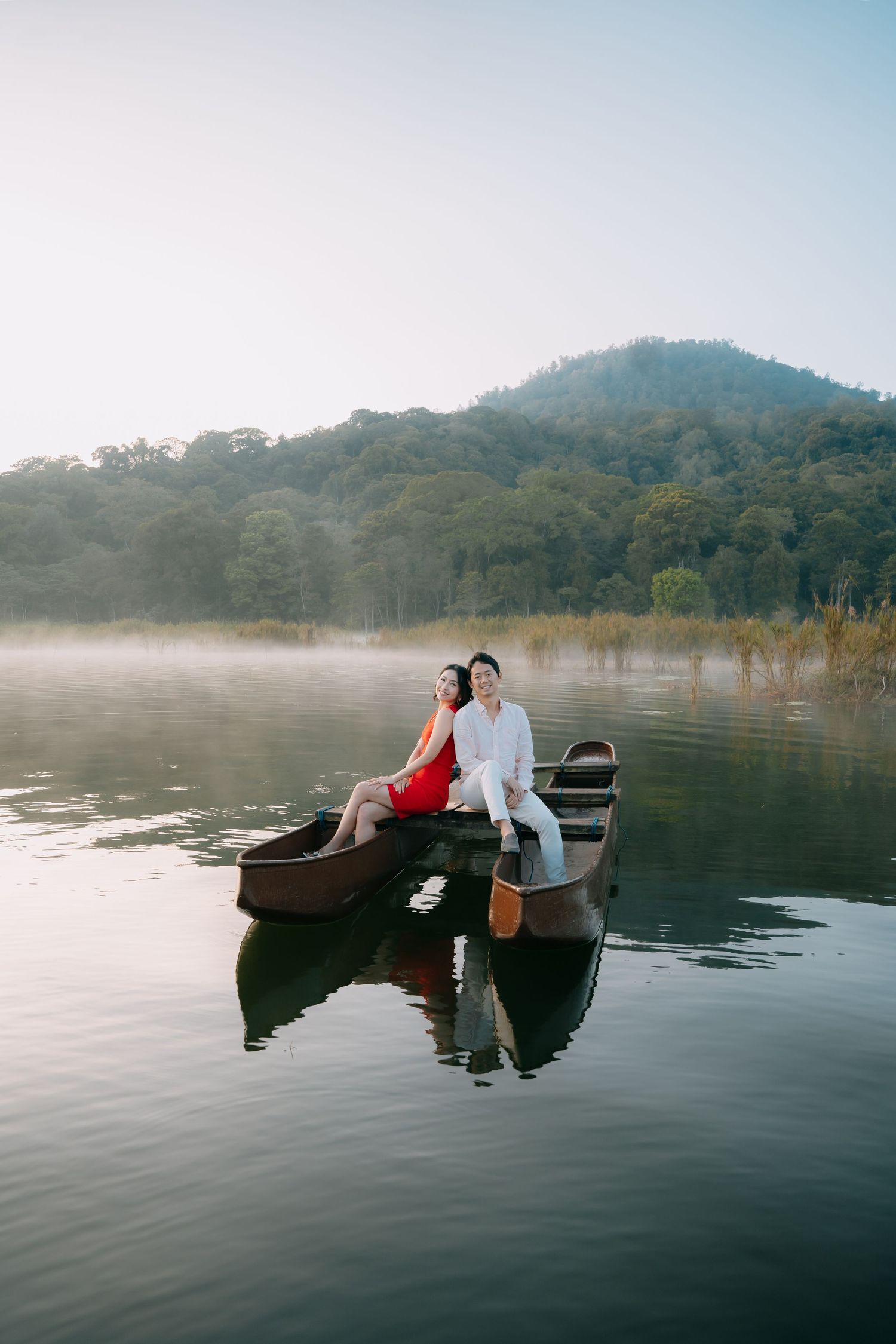 Two people in a wooden boat on a misty lake surrounded by forested hills during a tranquil morning.