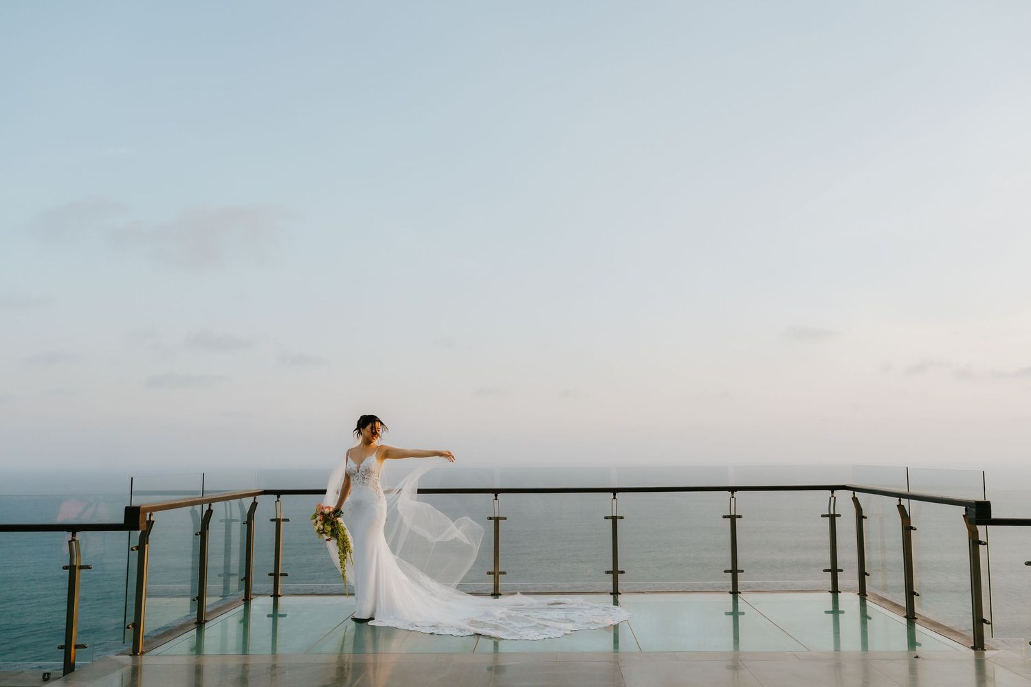 Bride in white gown poses on glass balcony overlooking ocean at sunset, veil flowing in the breeze.