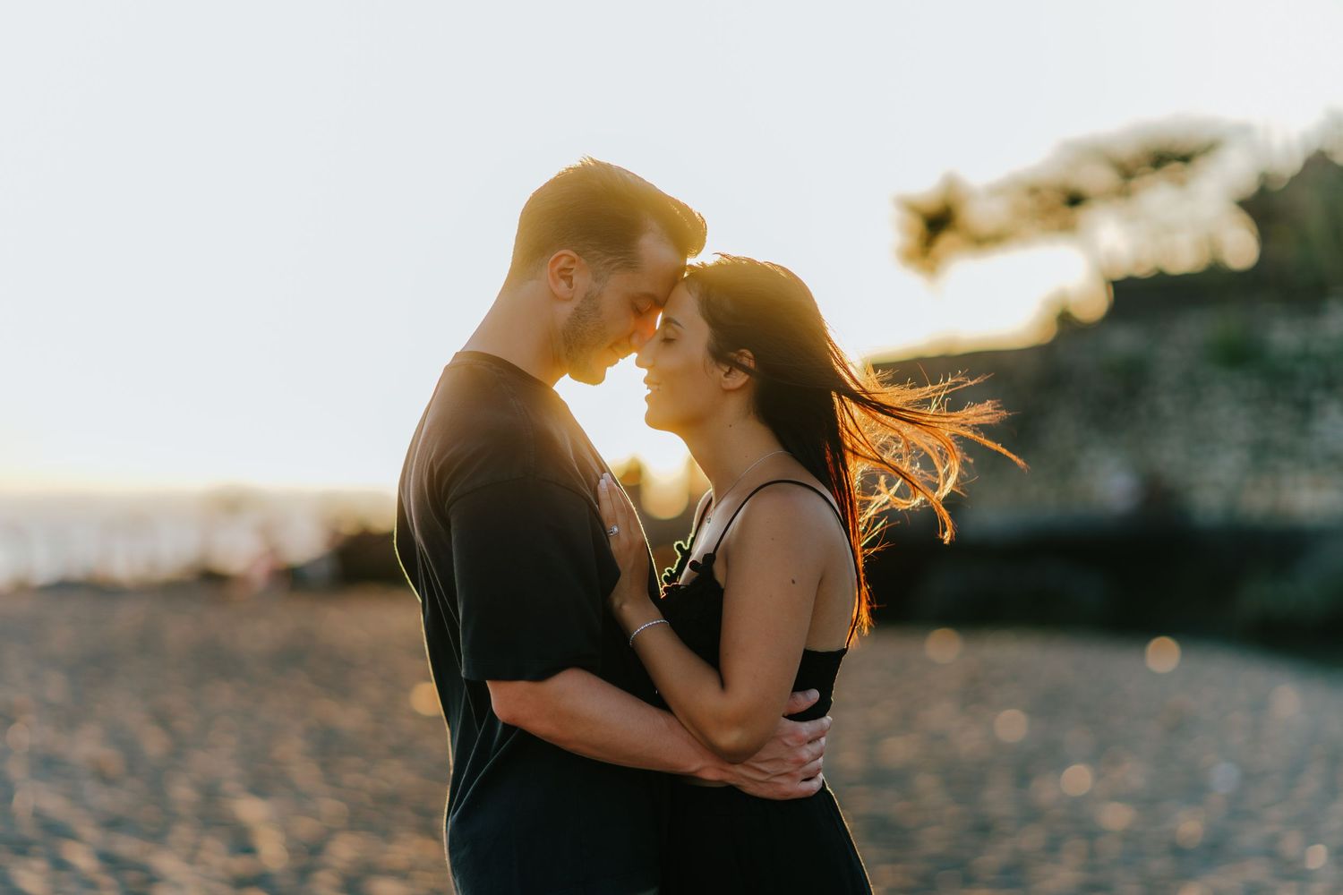 Couple embracing on beach at golden hour with sun flare in background.