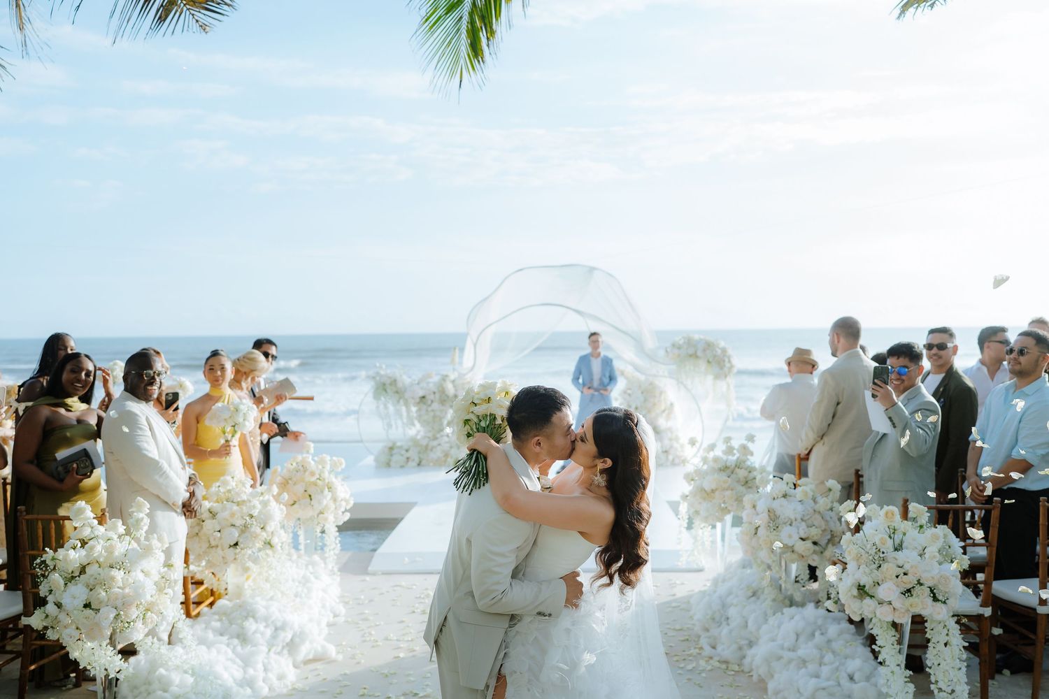 Couple shares a kiss at an elegant beachfront wedding ceremony with white floral arrangements and ocean backdrop.