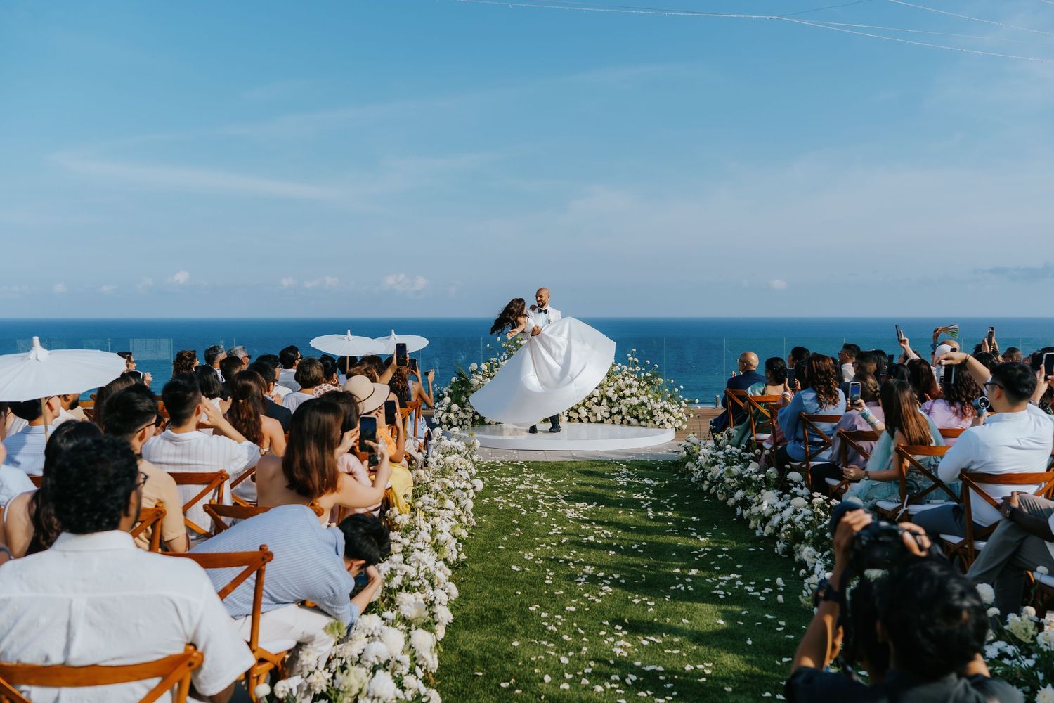 Outdoor cliffside wedding ceremony with couple kissing at altar, white floral aisle, ocean backdrop, guests seated on both sides.