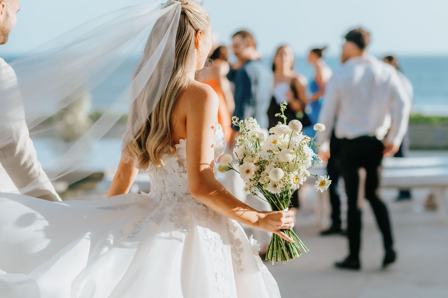 Bride in white wedding gown holding white floral bouquet with veil flowing, guests mingling in background at outdoor ceremony.