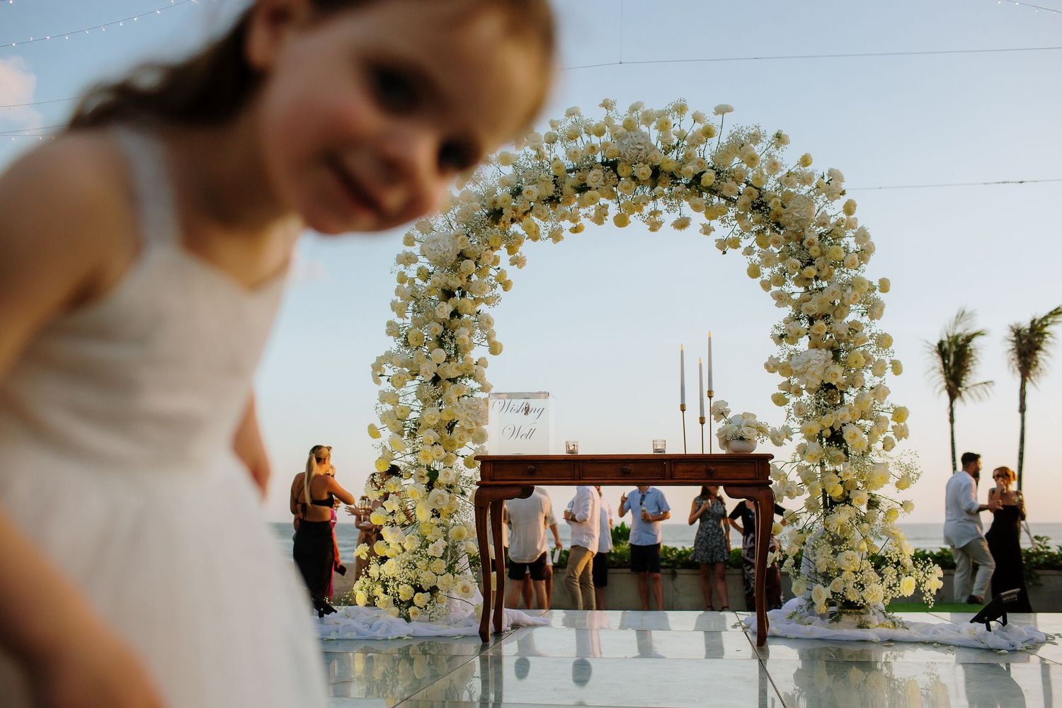Young girl smiles at outdoor wedding ceremony with elegant white floral arch and guests in background.