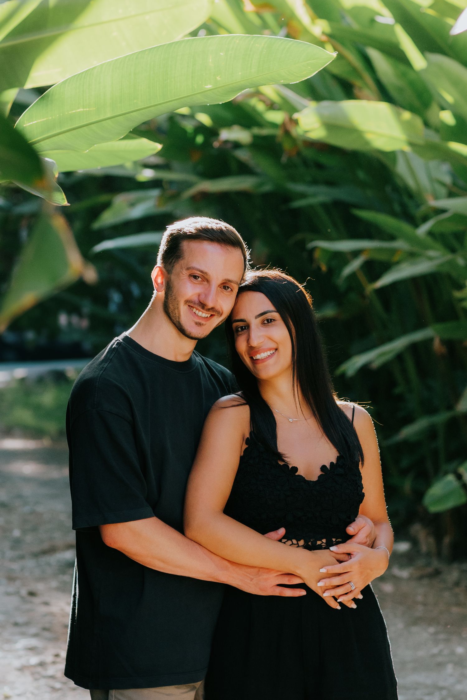 Happy couple in black attire posing together against a backdrop of large green leaves.