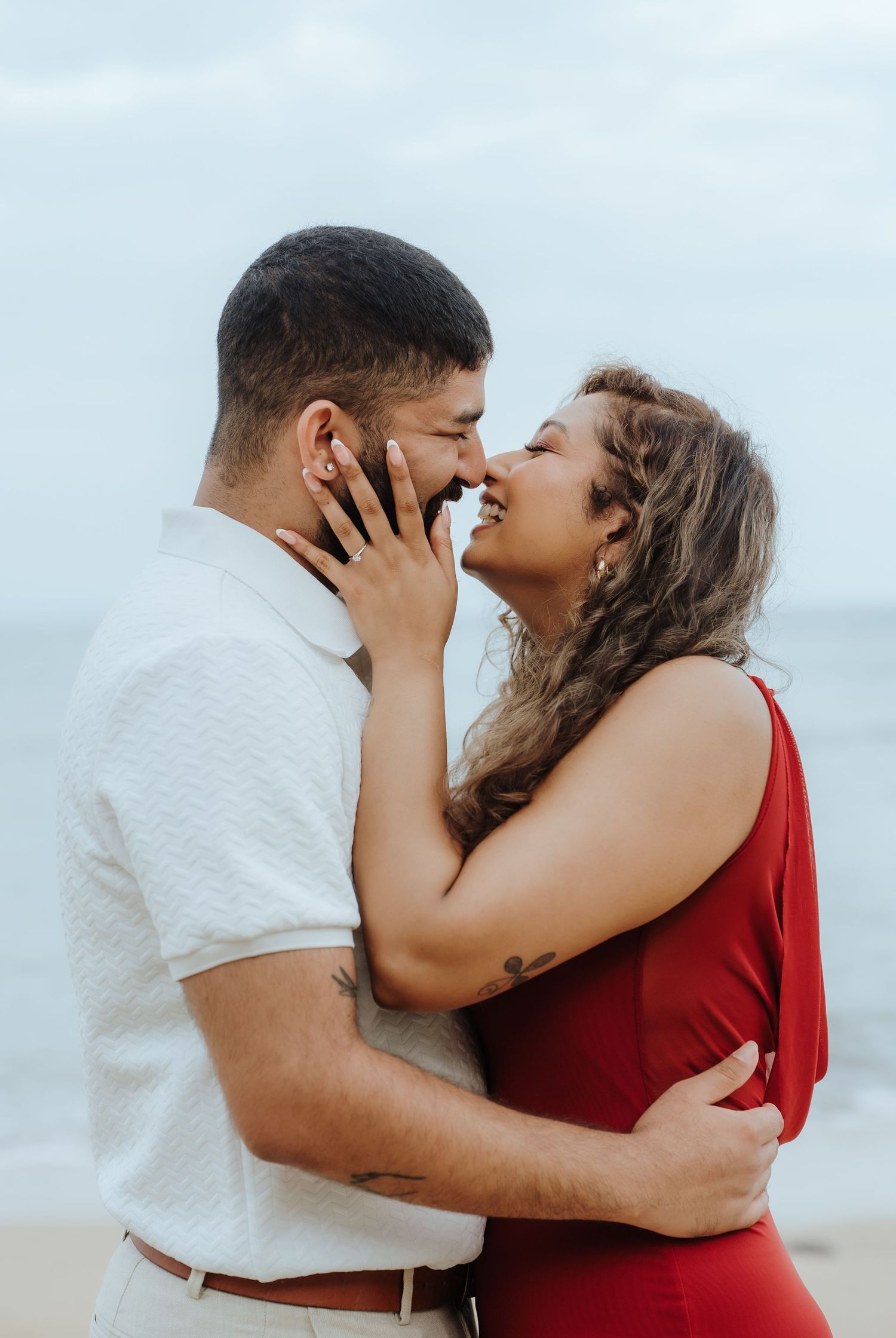 A romantic couple in red and white outfits embrace and kiss on a cloudy beach in a series of intimate photos.