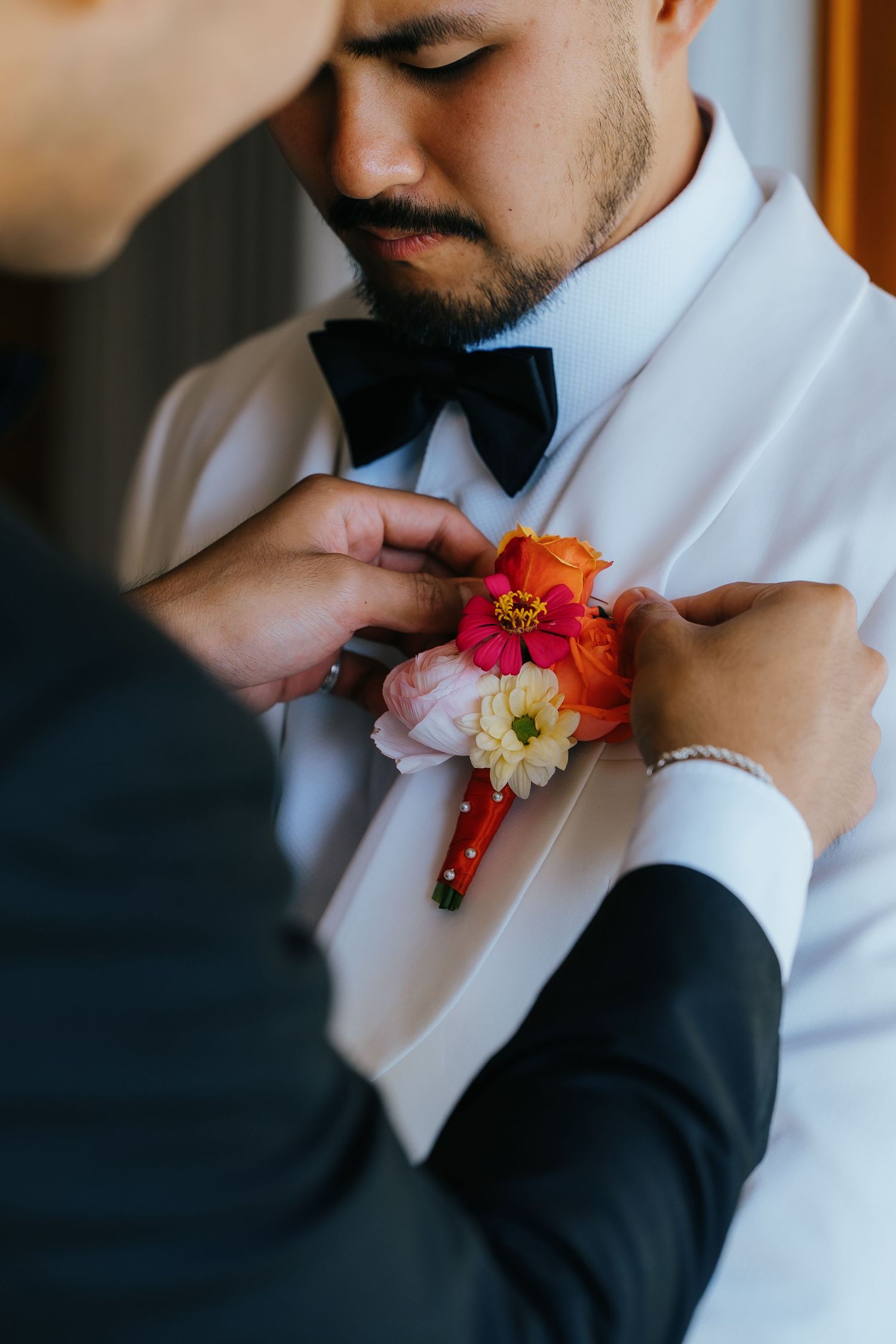 A groom in a white tuxedo and black bow tie has a colorful floral boutonniere pinned to his lapel.