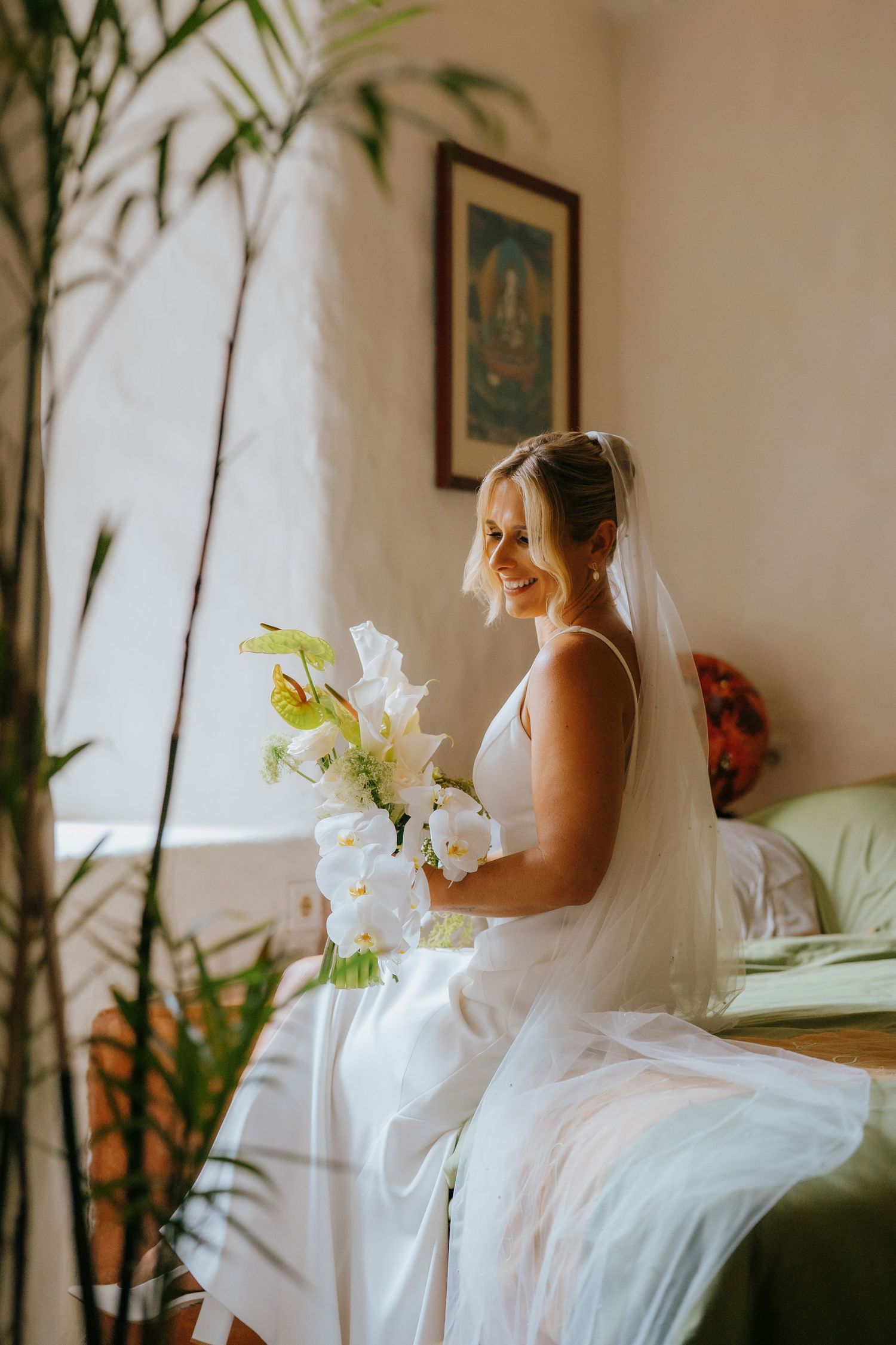 Smiling bride in white gown and veil holds bouquet of white flowers while sitting in a sunlit room.