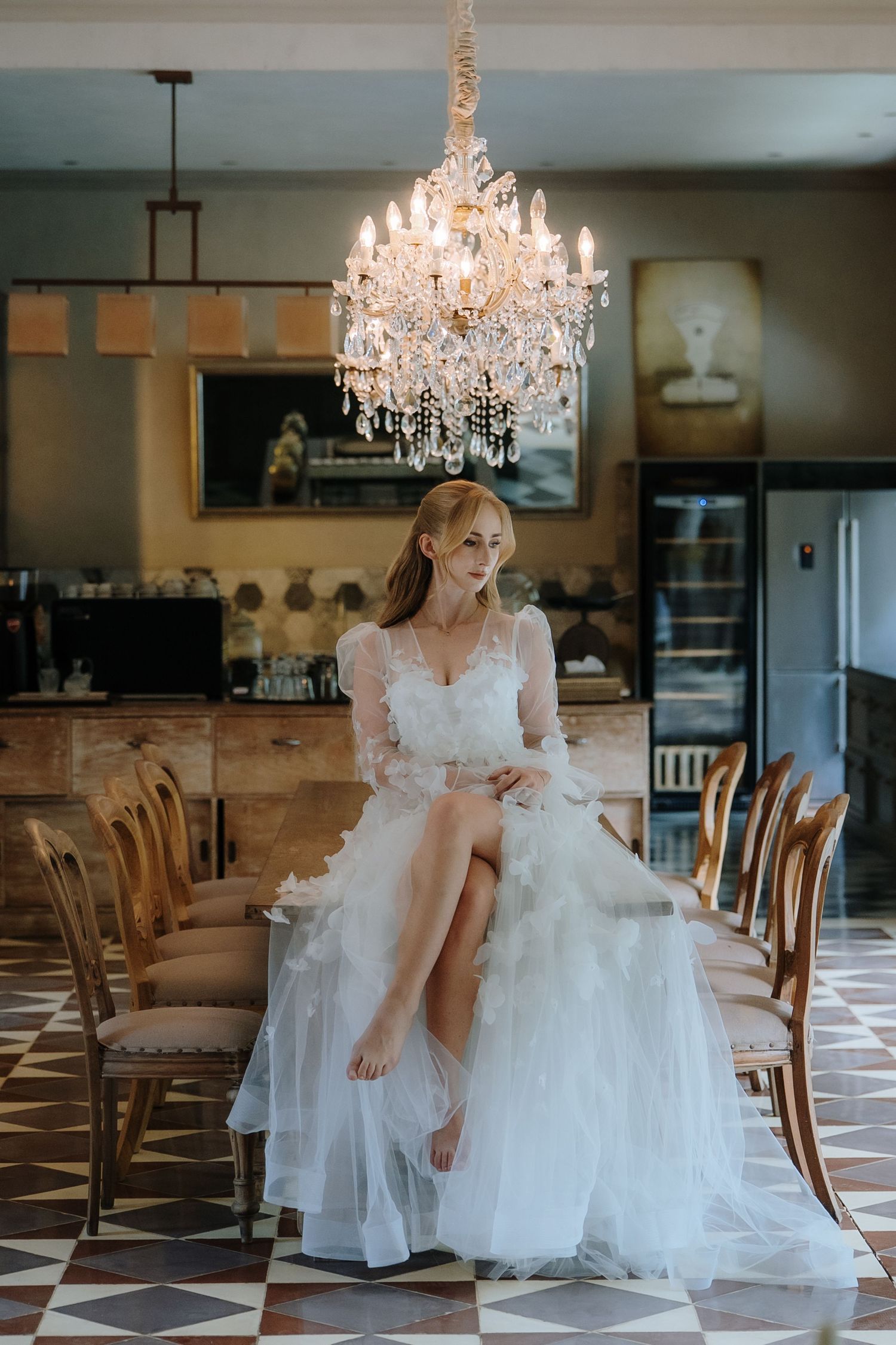 Bride in white lace gown seated on dining table under crystal chandelier in elegant restaurant setting.