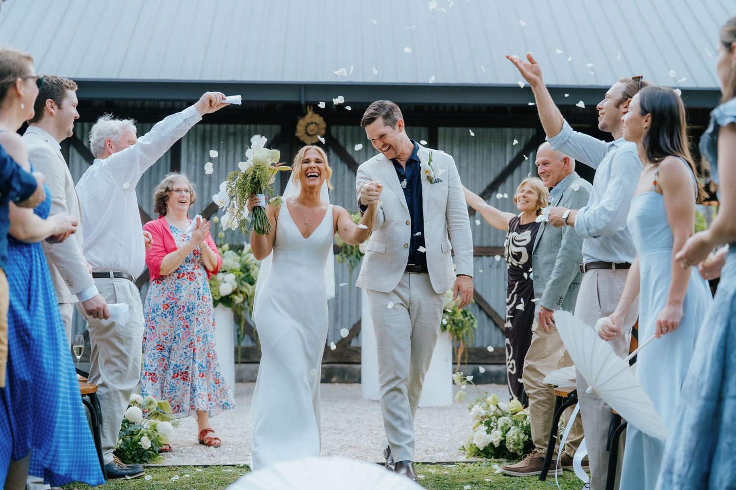 Bride and groom walk through confetti shower surrounded by cheering guests outside a barn wedding venue.
