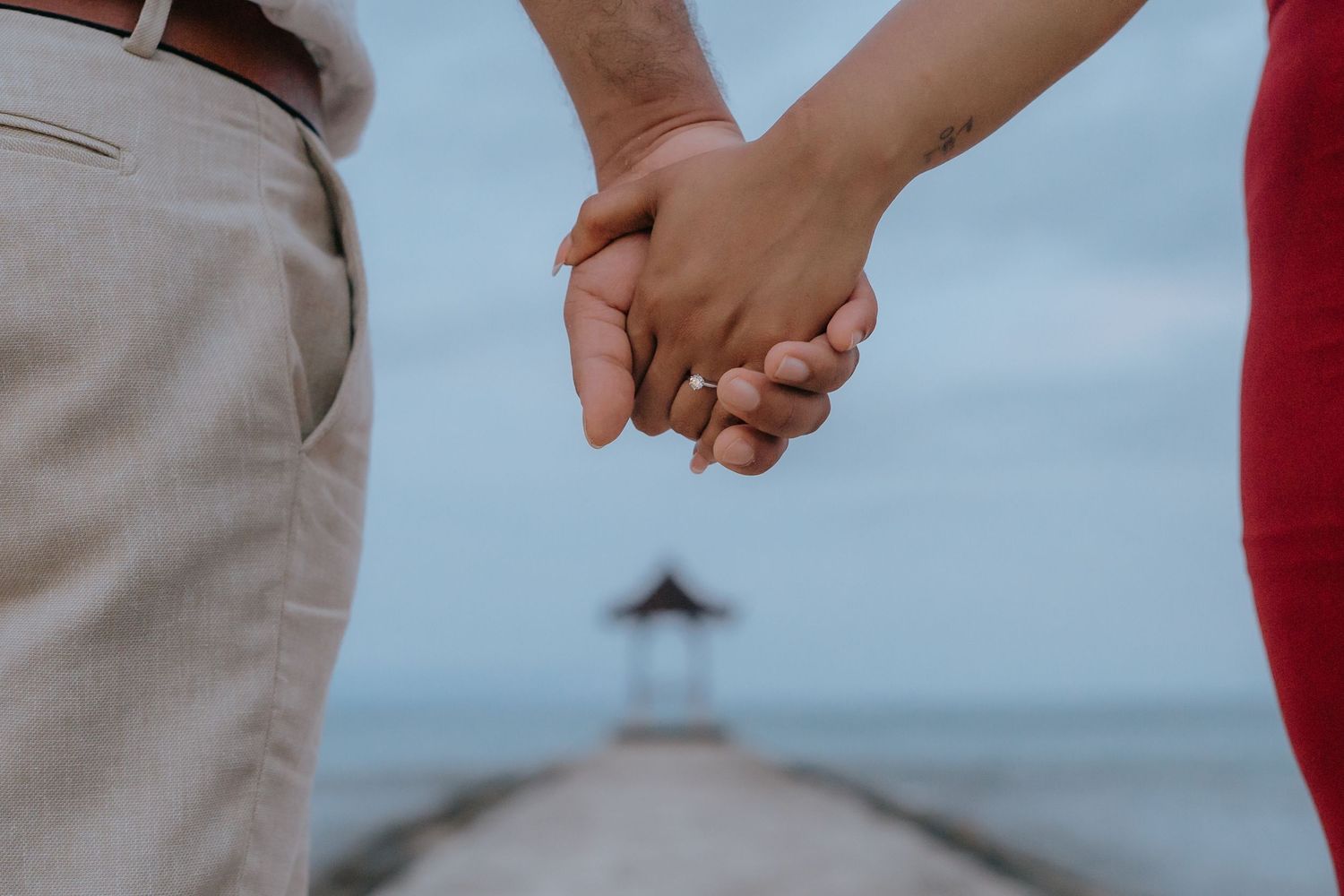 Two people hold hands while walking together on a beach during sunset with ocean waves in the background.